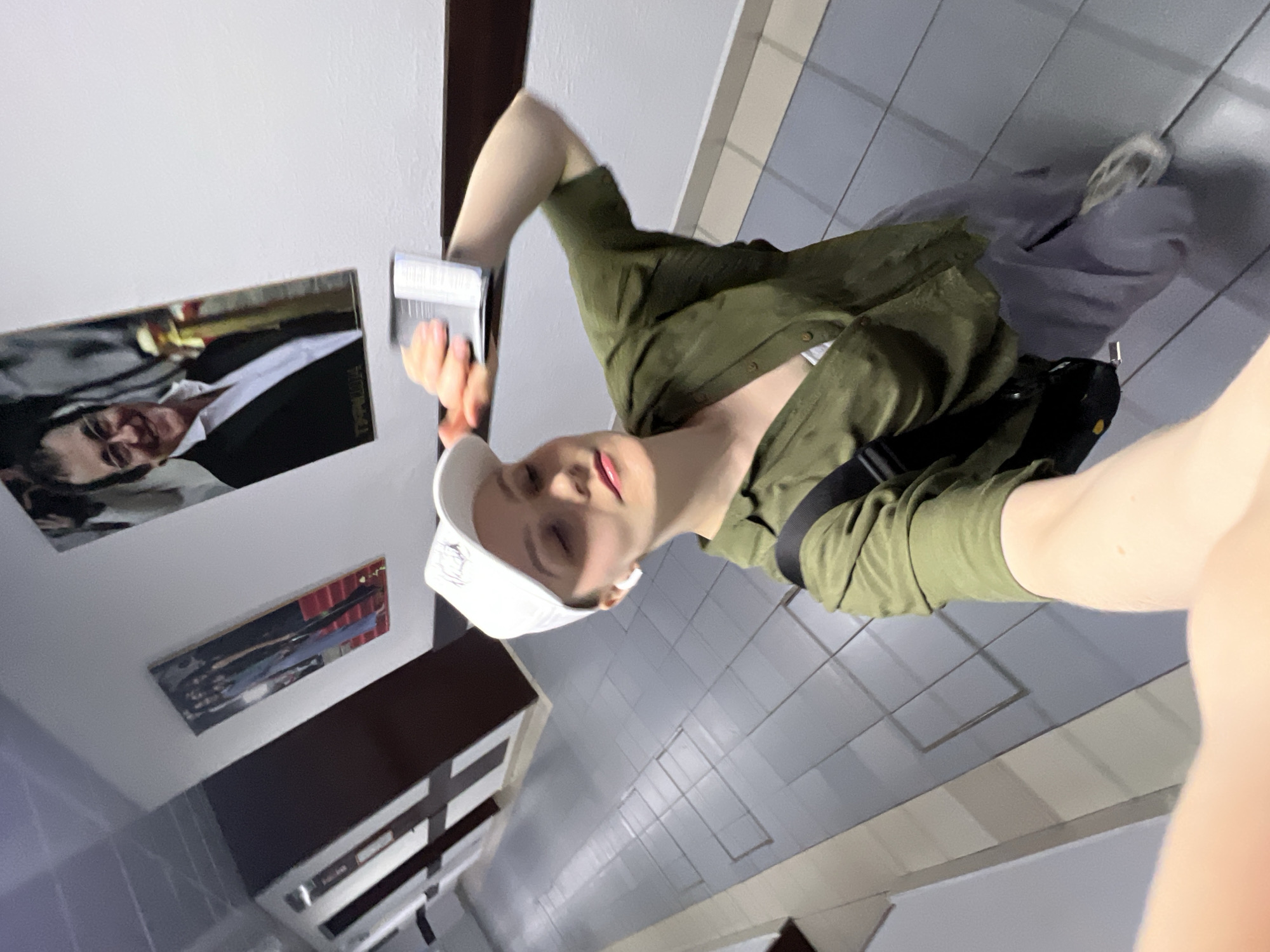 A person wearing an olive green shirt and a white baseball cap is taking a selfie in a building. They are holding a small rectangular object in their right hand. Behind them, there is a wall with two framed pictures and a dark cabinet. The building has a tiled floor and ceiling. The image was taken in Khimki, Russia.