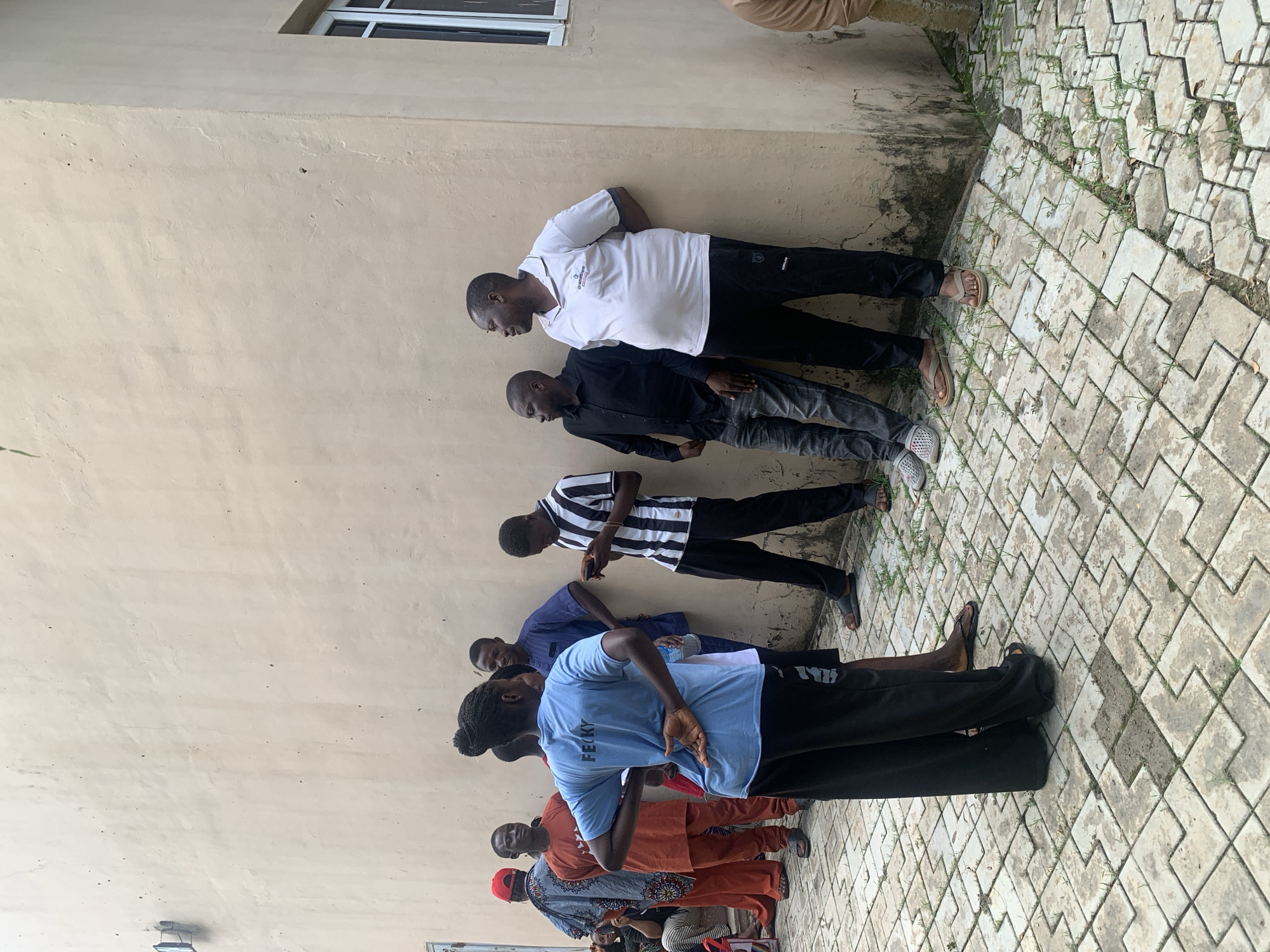 A group of eight people, appearing to be adults, are standing outdoors near a light beige building with a white-framed window. The ground is paved with interlocking gray and white concrete tiles, with some patches of grass growing between them.
From left to right:
- A person wearing a red cap and a dark blue and white patterned garment is standing on the far left.
- Next to them is a person in an orange outfit.
- A woman in a light blue t-shirt with "FECKY" printed on the back is the most prominent figure, facing away from the camera with her left hand behind her back. She is wearing black pants.
- A person in a dark blue tunic and dark pants stands behind her.
- A man in a black shirt and gray jeans is looking to his left.
- A man in a white polo shirt with a dark collar and logo, and black pants, is looking to his right.
- Further to the right, partially visible, is a person wearing a black and white striped long-sleeved shirt and black pants. They are looking down at a mobile phone.
- On the far right, a person in a light brown garment is partially visible, facing away from the camera.
The scene appears to be a casual gathering or waiting area. The building's wall has some dark staining and what appears to be a light fixture on the left side. The overall environment suggests an urban or semi-urban setting, possibly in Akwanga, Nigeria, given the typical architecture and clothing styles. The lighting suggests it is daytime.
