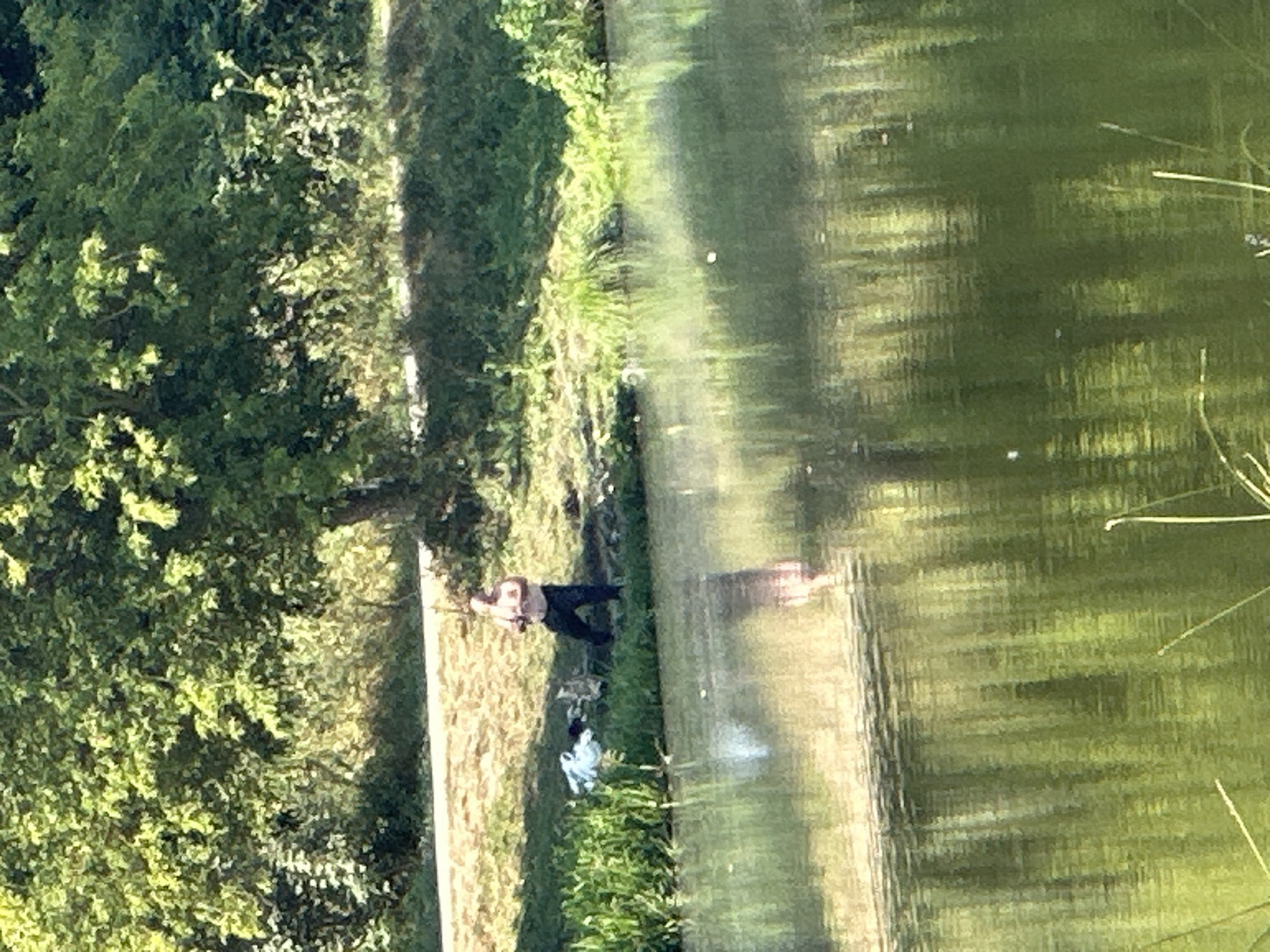 A man is standing on the bank of a body of water, near a tree. He is wearing a light-colored shirt and dark pants. There is a pile of white clothing to his left. The water has reflections of trees and the sky. The location is the city of Makiv, Ukraine.