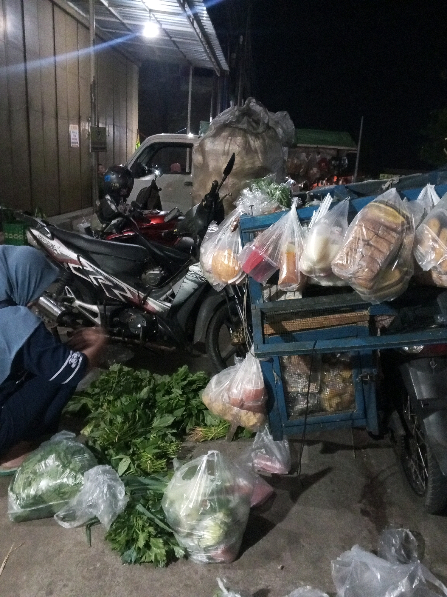 The image depicts a street scene at night in Gentan, Indonesia. In the foreground, several bags of fresh produce, including leafy green vegetables and what appears to be celery, are arranged on the ground. A person wearing a blue hijab and dark blue pants is bending down, interacting with the vegetables. A motorcycle with a black and red body is parked nearby, with a black helmet on its seat. Behind the motorcycle, a blue metal cart is filled with various items bagged in plastic, including what looks like bread loaves, bottles, and other food products. A white pickup truck is visible in the background, along with a large pile of what appears to be fabric or debris. The area is lit by artificial lighting, casting shadows on the ground. The scene suggests a vendor or a market setting.