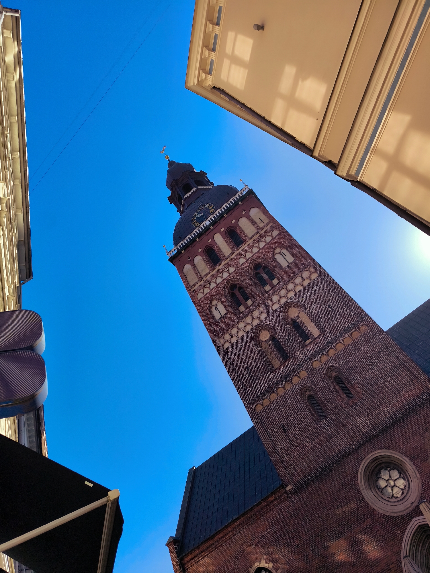 A low-angle, upward shot captures the imposing brick bell tower of Riga Cathedral against a clear blue sky. The tower, constructed from reddish-brown bricks, features numerous arched windows with decorative detailing and a clock face near its apex. The roof of an adjacent building, made of dark material, is visible to the right of the tower. The left side of the frame is occupied by the facades of two buildings, one pale yellow with visible window reflections and another with a dark awning. The scene is set in Riga, Latvia.