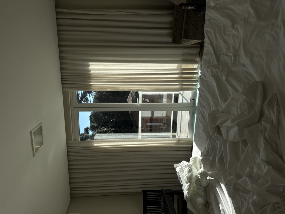 The image displays the interior of a room, located in Canela, Brazil. A large bed in the foreground is covered with disheveled white sheets and a duvet. Strong natural light enters through a tall, white-framed glass double door, positioned centrally in the background, which leads to an outdoor tiled balcony. Floor-to-ceiling light-colored curtains are partially drawn back on either side of the glass door. To the left of the bed, a dark wooden chair is partially visible. To the right, a light-colored armchair with a dark bottle on an adjacent surface is present. Beyond the balcony, a red-brick building with a brown roof is visible, partially obscured by dense, dark green trees. A clear blue sky is visible above the trees. The room has a light-colored ceiling with a square, recessed light fixture. No individuals are present in the scene.