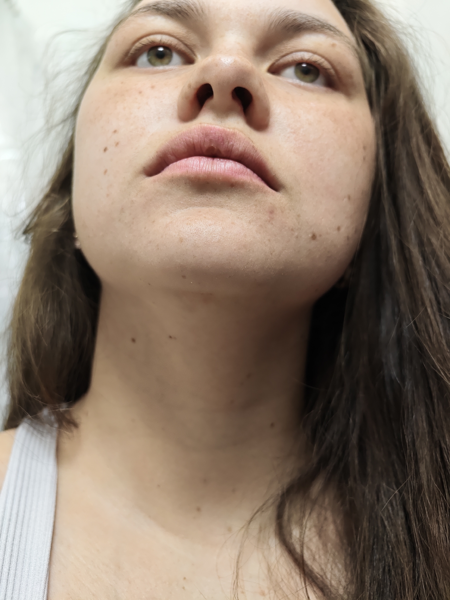 A close-up, upward-angle photograph depicts a woman's face, neck, and upper chest. She has light skin with freckles, brown hair, and light-colored eyes. The background appears to be a plain, light-colored wall. The individual is wearing a light-colored tank top. The photograph's location in Chaikovsky, Russia, is not evident from the image.