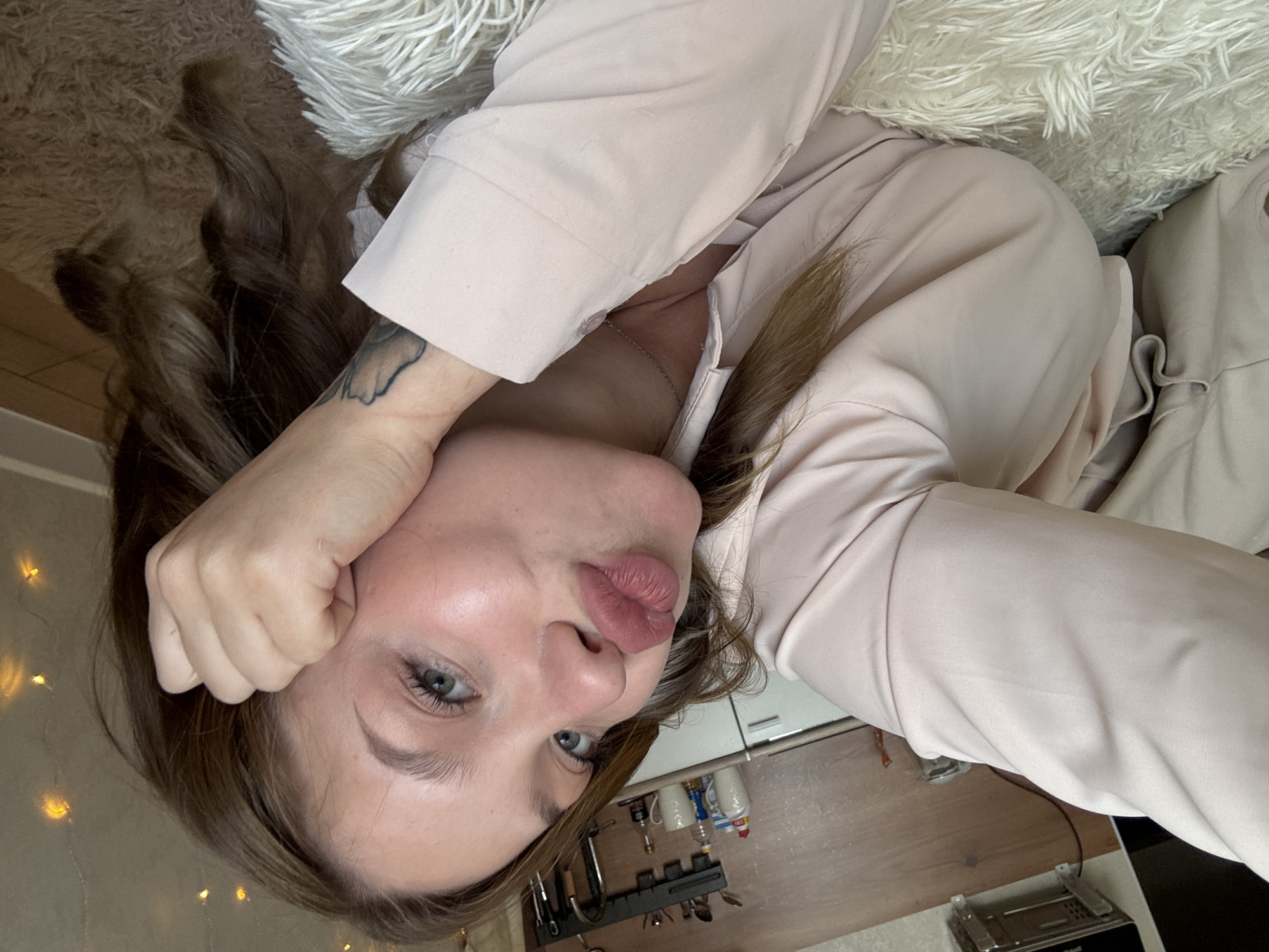 A woman is lying on a furry rug, facing the camera, with her head resting on her hand. She has her lips pursed in a kissing gesture. She is wearing a light pink collared shirt. Her hair is long and brown. In the background, there are kitchen cabinets and a shelf with items on it. There are also some lights that appear to be fairy lights. The overall lighting is dim, with some warm light from the fairy lights.
