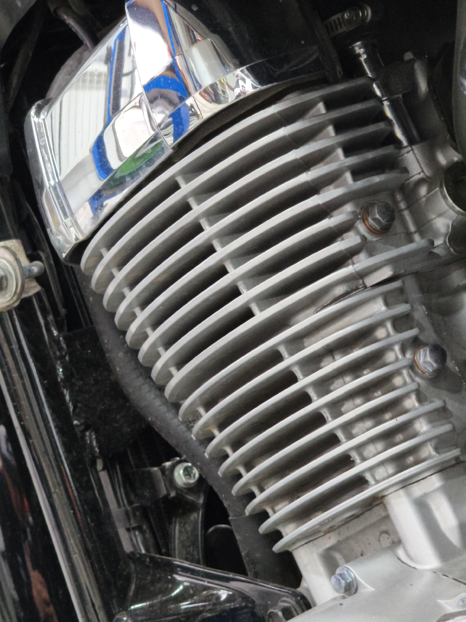The image shows a close-up view of a motorcycle engine, focusing on the cylinder fins and a chrome cover. The engine appears to be metallic and has a ribbed texture. There are several bolts and hoses visible. The background is dark and out of focus, suggesting it's likely within a workshop or garage. There are no people or any indication of an outdoor or urban environment. The location in the city of Noviy, Russia is not discernible from the image itself.