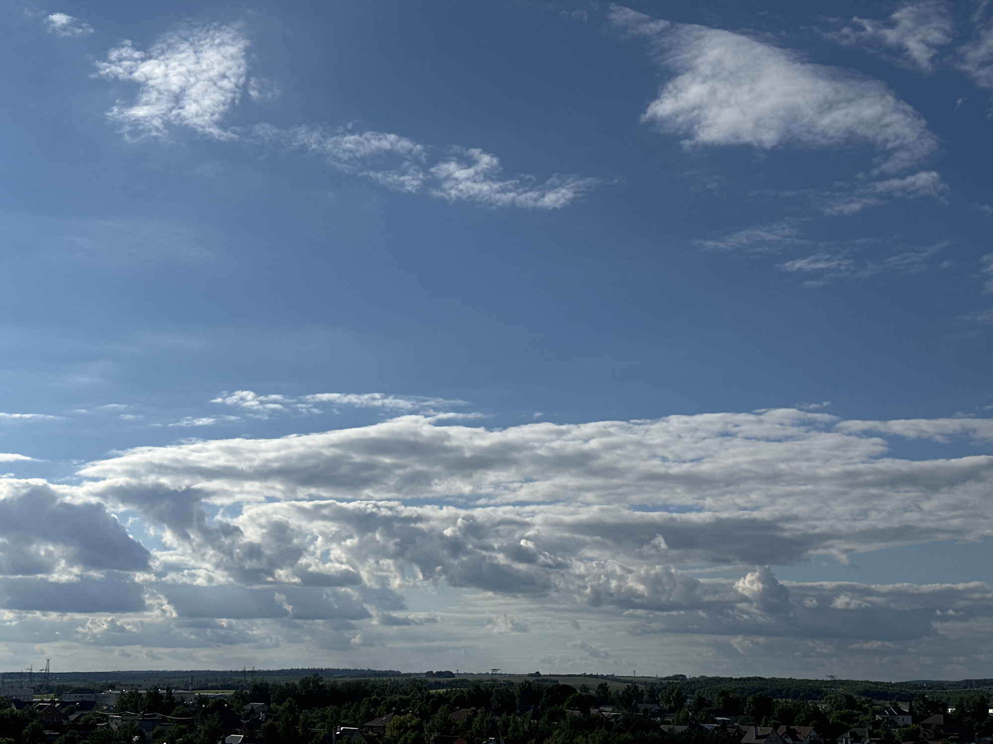 This is a wide-angle photograph of a blue sky with scattered clouds, taken from a high vantage point overlooking a rural landscape. The sky takes up the majority of the frame, with a band of dense, layered clouds stretching across the mid-ground. These clouds are a mix of white and grey, indicating varying thicknesses and possibly some precipitation. Lighter, wispy clouds are visible higher in the sky.
Below the clouds, a dark green expanse of trees forms the horizon line, suggesting a forested or heavily wooded area. Nestled within the trees are rooftops of houses, indicating a small village or suburban settlement. The scene appears to be set in a rural or semi-rural location. The lighting suggests it is daytime, possibly late afternoon, as the light is somewhat diffused by the clouds but still bright. There are no visible people or specific objects that dominate the scene. The overall mood is peaceful and expansive, characteristic of a natural landscape.
The location context is provided as Шчомысліцкі сельскі Савет, Belarus. No text is visible in the image.