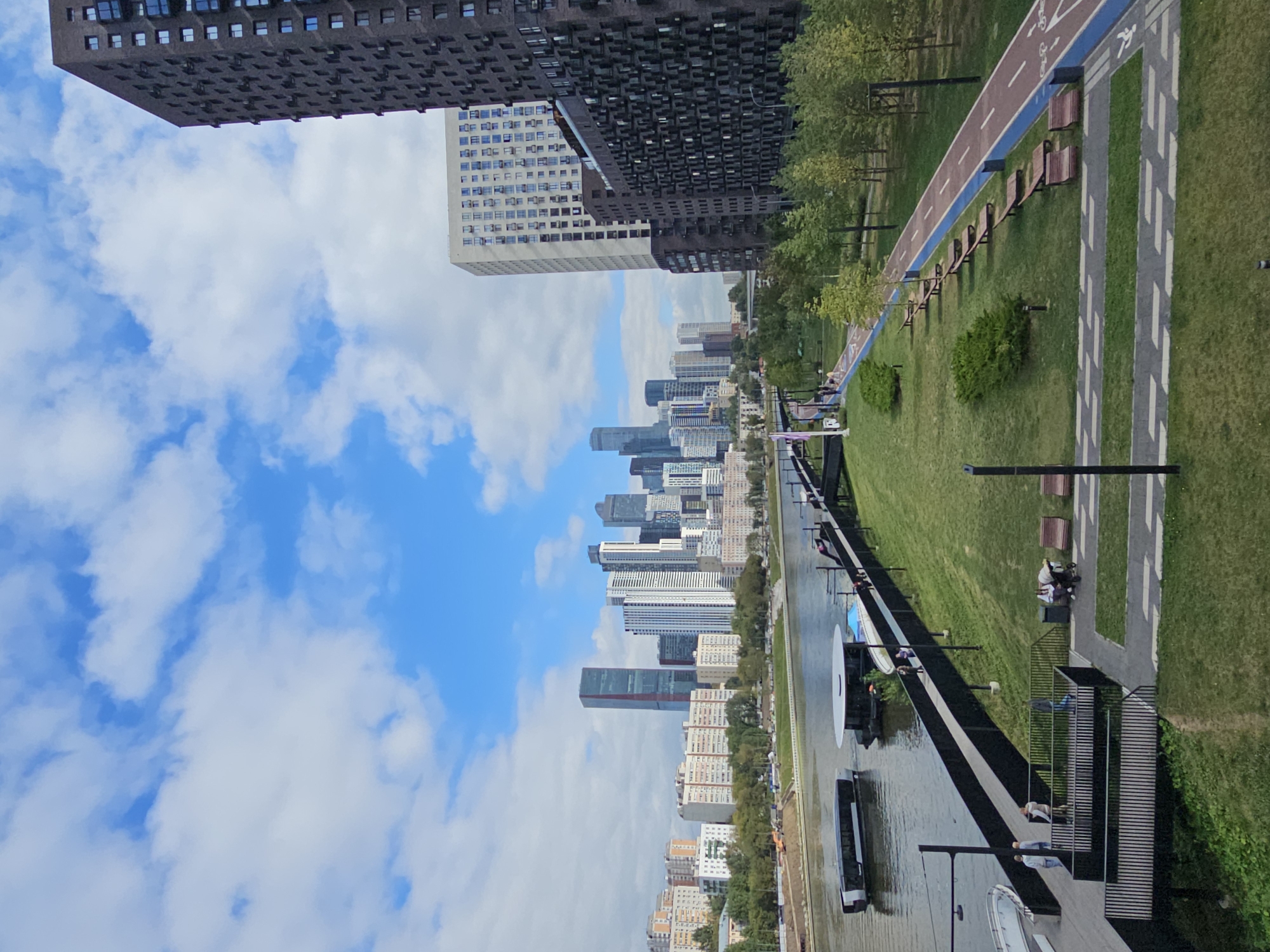 The image depicts a modern urban park situated along the Moscow River in Moscow, Russia. In the background, the Moscow City skyline is visible, featuring numerous contemporary skyscrapers, including the Neva Towers and the Federation Tower. In the foreground, a well-maintained park area includes a paved walkway, a dedicated bike path with a red surface and blue borders, and several benches. Several trees and green spaces are also present. On the Moscow River, a boat is seen in the water, and a floating structure, possibly a cafe or restaurant, is docked. The sky is partially cloudy with blue patches. Several individuals are present in the park, some walking along the walkway and others seated on benches. The scene suggests a recreational and residential area within the city.
