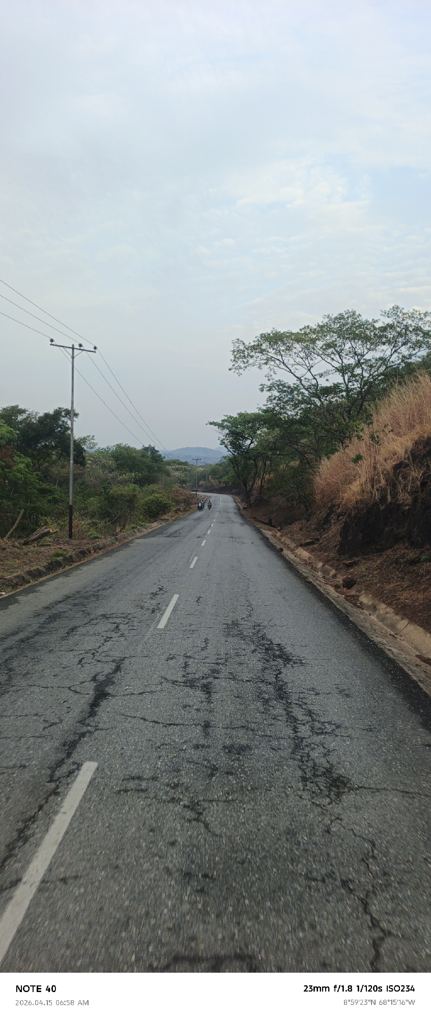 The image depicts a cracked asphalt road stretching into the distance, flanked by vegetation and telephone poles. Two figures on motorcycles are visible further down the road. The sky is overcast. According to the metadata, the image was taken on April 15, 2026, at 6:58 AM, and the location is 8°59'23"N, 68°15'16"W. This places the location near La Cruz, Venezuela.