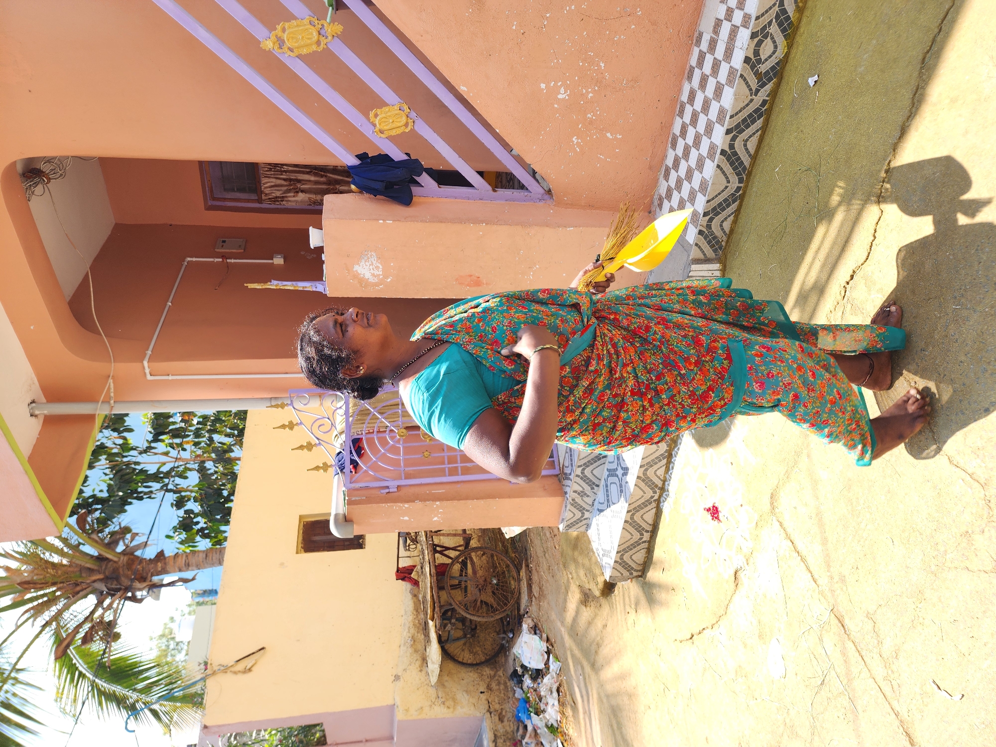 An adult woman is depicted in an outdoor residential area of Pedda Kammavaripalli, India, under clear, sunny conditions. The woman, barefoot, is shown in profile, facing right. She has dark skin, dark hair tied back, and wears a teal-colored blouse beneath a vibrant floral sari with red, orange, green, and teal patterns. She is adorned with a gold-colored bangle on her right wrist and a dark beaded necklace, with a small stud in her right nostril. Her left hand holds a yellow plastic dustpan containing a natural fiber broom.
The immediate ground is a cracked, light-colored concrete or paved surface, exhibiting distinct shadows. To the woman's right, a peach-colored house features a tiled base with a black and white geometric pattern, and purple stairs/railing with two ornate golden embellishments. A dark blue garment hangs over a structural pillar of the house. An arched entrance leads into a darker interior with a brown curtain, and exposed electrical wiring is visible overhead.
In the midground to the left, another faded peach-colored building has a small, dark window. Adjacent to this building, a pile of assorted refuse (plastic, paper) accumulates next to a rustic, two-wheeled cart. Above this structure, a large palm tree with visible coconuts stands against a bright blue sky. White PVC piping runs horizontally on the walls of both houses.