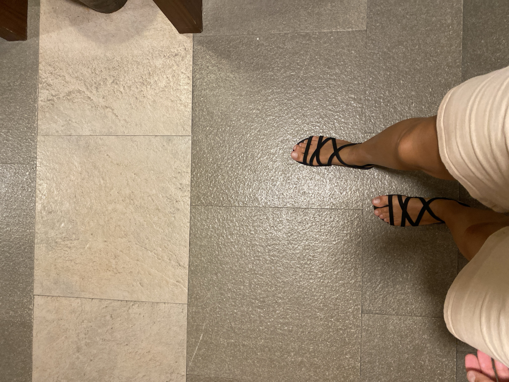 A high-angle shot shows a person's legs and feet from the knees down, standing on a tiled floor. The person is wearing beige shorts and black strappy sandals. The floor is made of two different types of tiles: large, light-colored, rough-textured tiles in the upper portion of the image, and smaller, dark gray, pebbly textured tiles in the lower portion. The transition between the two tile types creates a clear horizontal line. A dark wooden beam is visible on the right edge of the frame, suggesting the presence of furniture or an architectural element. The lighting is somewhat dim, casting a shadow from the person's right leg onto the tiles. There is no discernible indication of the city of Aksu, Turkey, within the image itself.