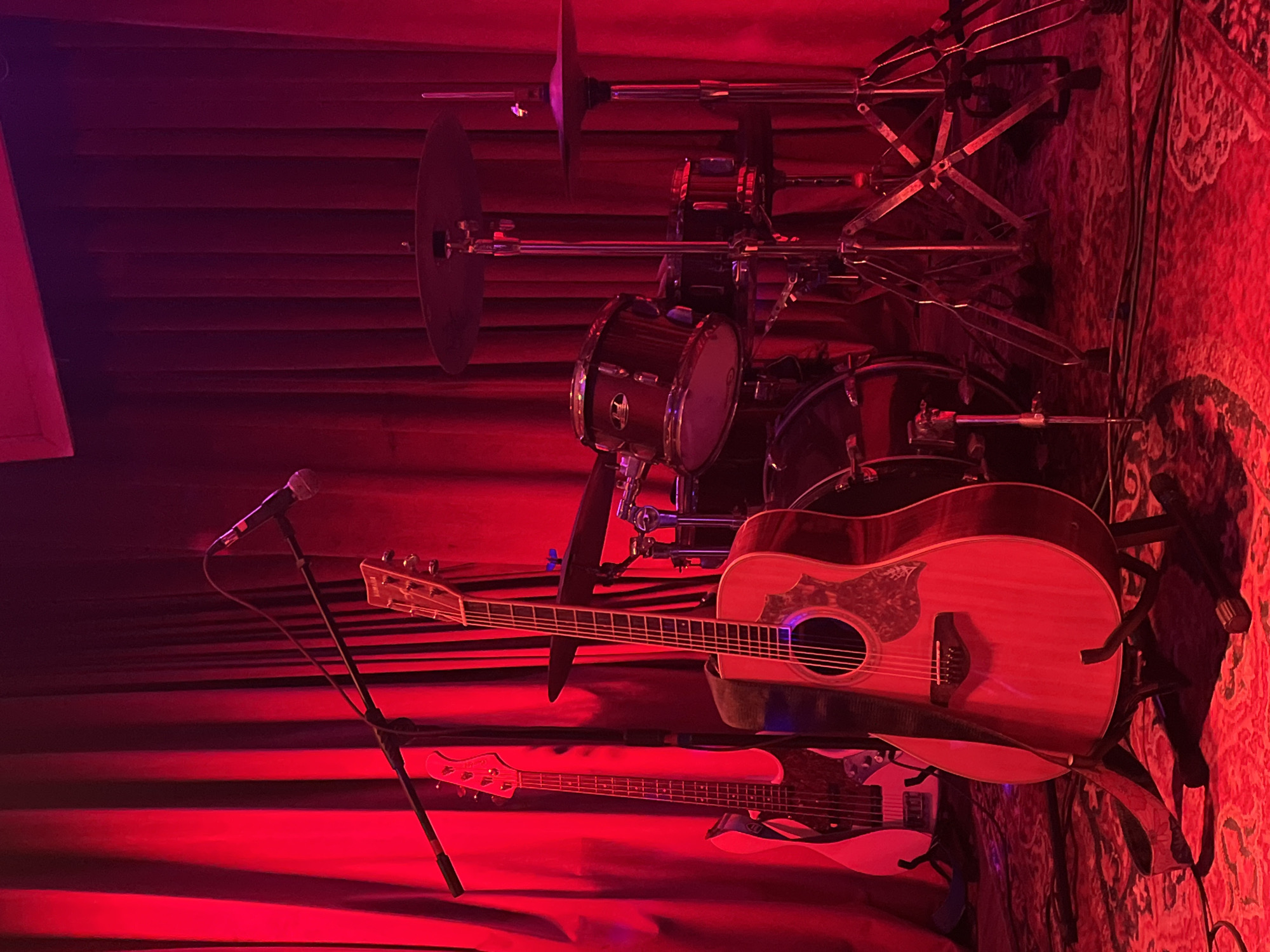 A dimly lit stage bathed in red light showcases musical instruments and equipment. An acoustic guitar rests on a stand in front of a drum set. A white bass guitar is visible below the acoustic guitar. A microphone stands to the left. The background is a red curtain. The location is likely a music venue, possibly in Đà Nẵng, Vietnam, given that it's in the request.