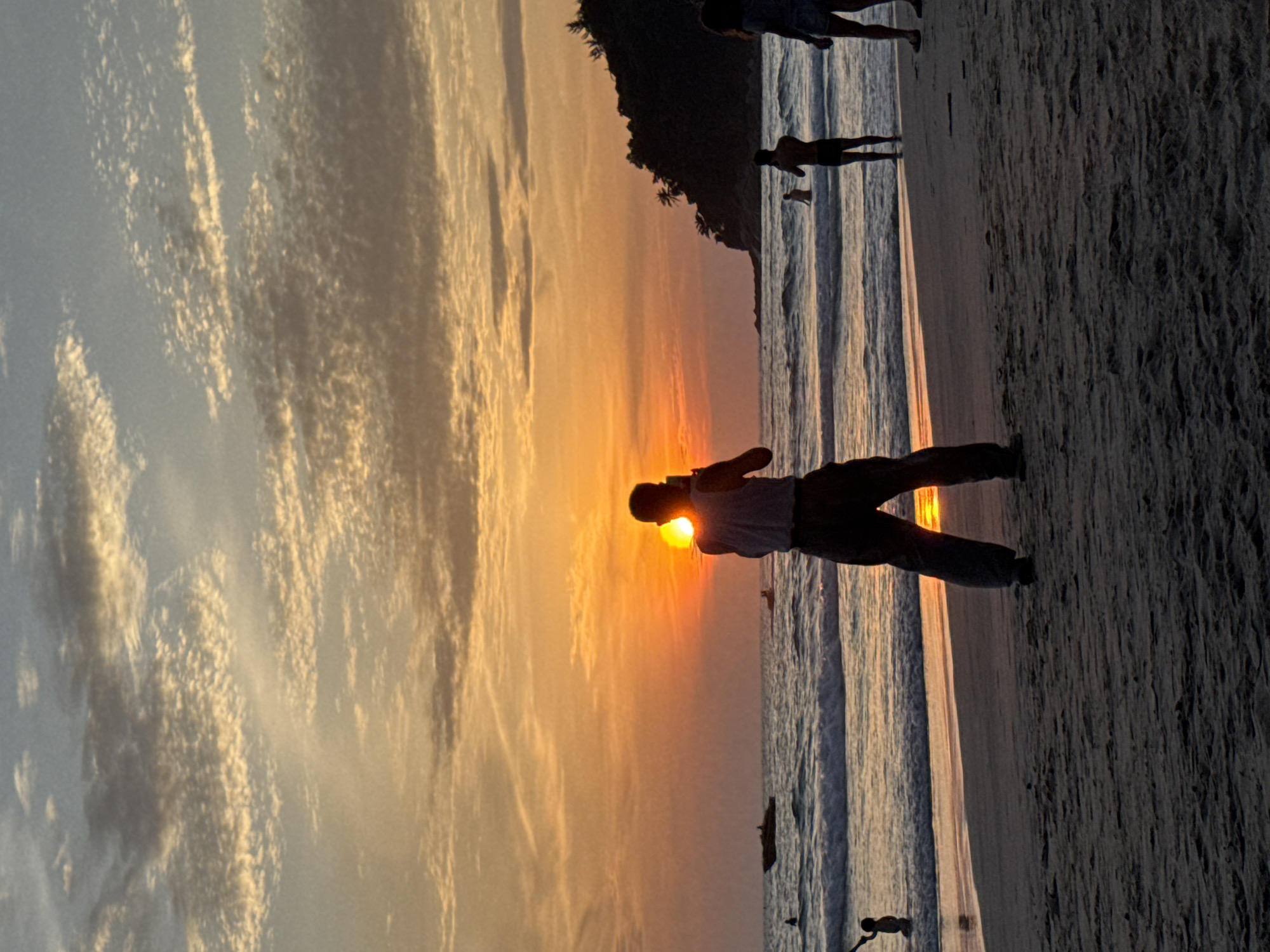 A silhouette of a person stands on a beach in Karon, Thailand, facing the setting sun. The sun is a bright orange disc low on the horizon, partially obscured by clouds. The person, dressed in a vest and pants, is holding a phone up to take a picture. Waves gently lap at the shore, and other people are visible in the water and on the sand. A boat is seen in the distance on the ocean. The sky is filled with streaks of orange, yellow, and blue. The beach is composed of dark sand. A forested hill rises in the background to the right.