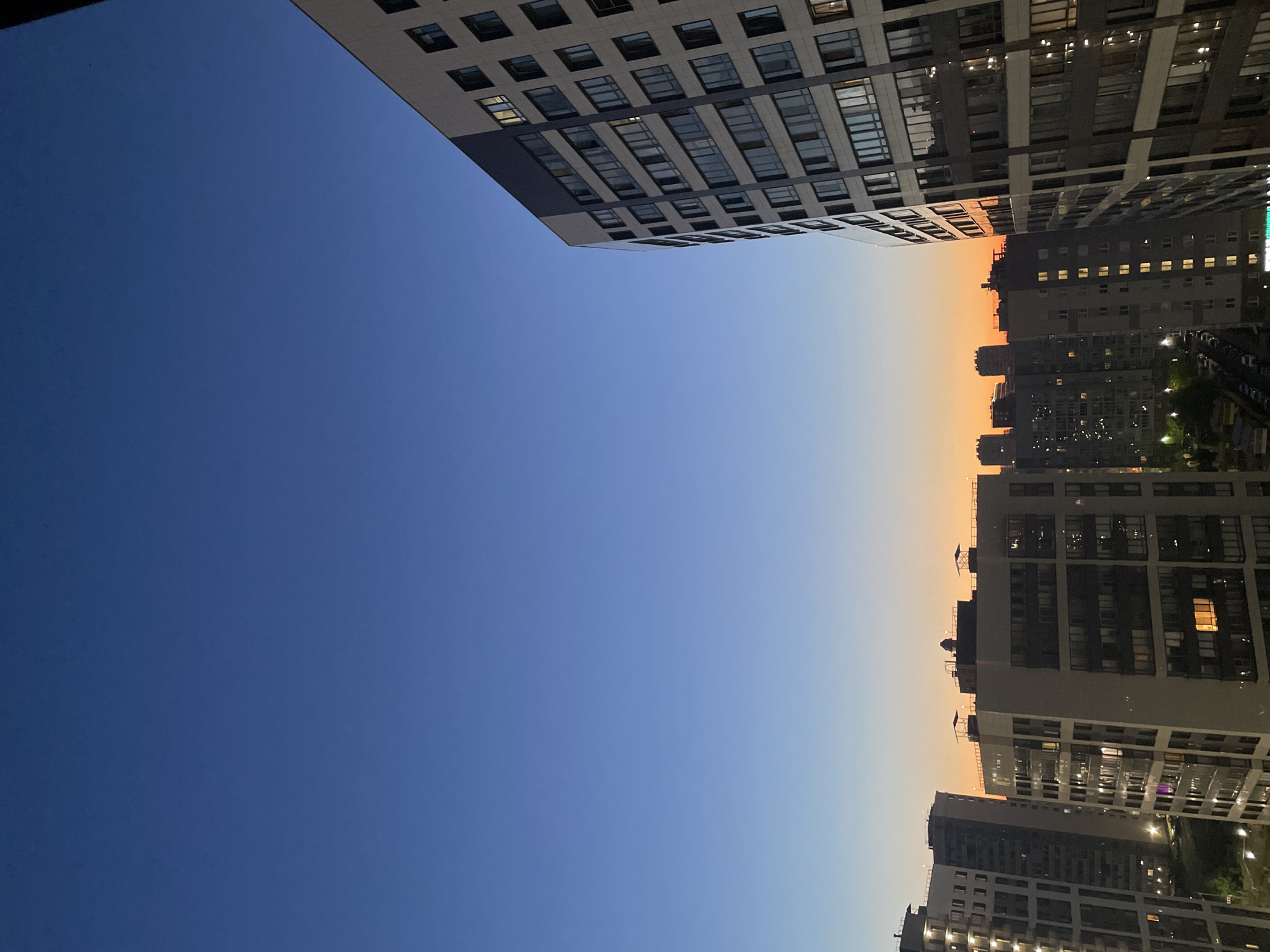 The image captures a twilight view of a residential area in Sinyavino, Russia. The sky displays a gradient of deep blue at the top transitioning to a soft orange and yellow hue near the horizon, indicating the sun has recently set. Silhouetted against the sky are several modern, multi-story apartment buildings. The building in the foreground, on the right side of the image, is tall and features a facade of windows and balconies. Other apartment buildings of varying heights and architectural styles are visible in the middle ground and background, with some windows illuminated, suggesting the presence of occupants. The overall scene depicts a typical urban evening environment.