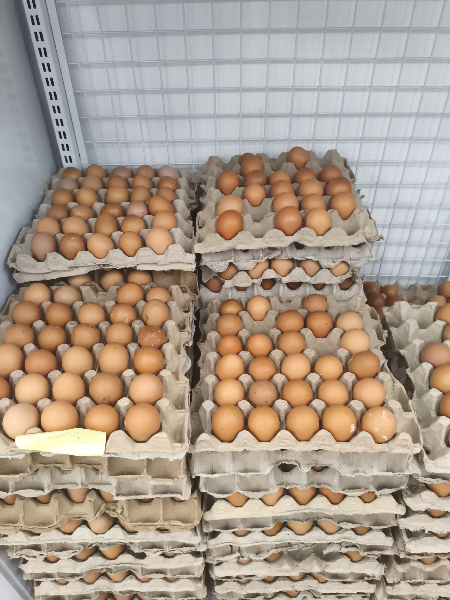 This is a close-up, eye-level shot of stacks of brown eggs in cardboard cartons, likely taken in a marketplace or grocery store in Depok, Indonesia. The eggs are mostly uniformly colored, with some variations in shade. They are arranged in rows within the cartons, with a yellow price tag visible on one of the cartons in the foreground. The background features a grid-like pattern, possibly the interior of a refrigerator or shelving unit. The lighting is even and bright, suggesting an indoor setting. There are no people or animals visible in the frame.