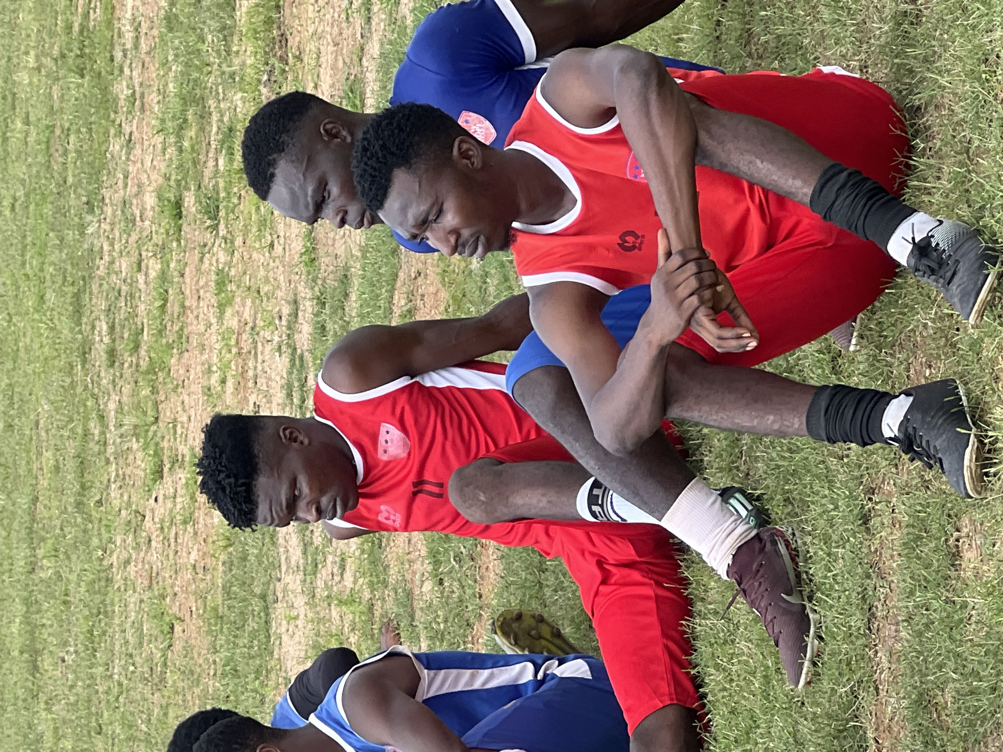 The image depicts a group of young men, likely a football team, resting on a grassy field in Jos, Nigeria. The men are dressed in athletic attire. Two men in the foreground are wearing red sleeveless jerseys and red shorts or pants. The man on the left is wearing white socks with black stripes and dark athletic shoes. The man on the right is wearing black socks and black athletic shoes. Both men are sitting with their legs extended. In the background, other individuals in blue jerseys and blue shorts are visible. One man in a red jersey with the number 11 is sitting with his legs bent, looking to the left. The environment appears to be a sports field or park with dry grass.
