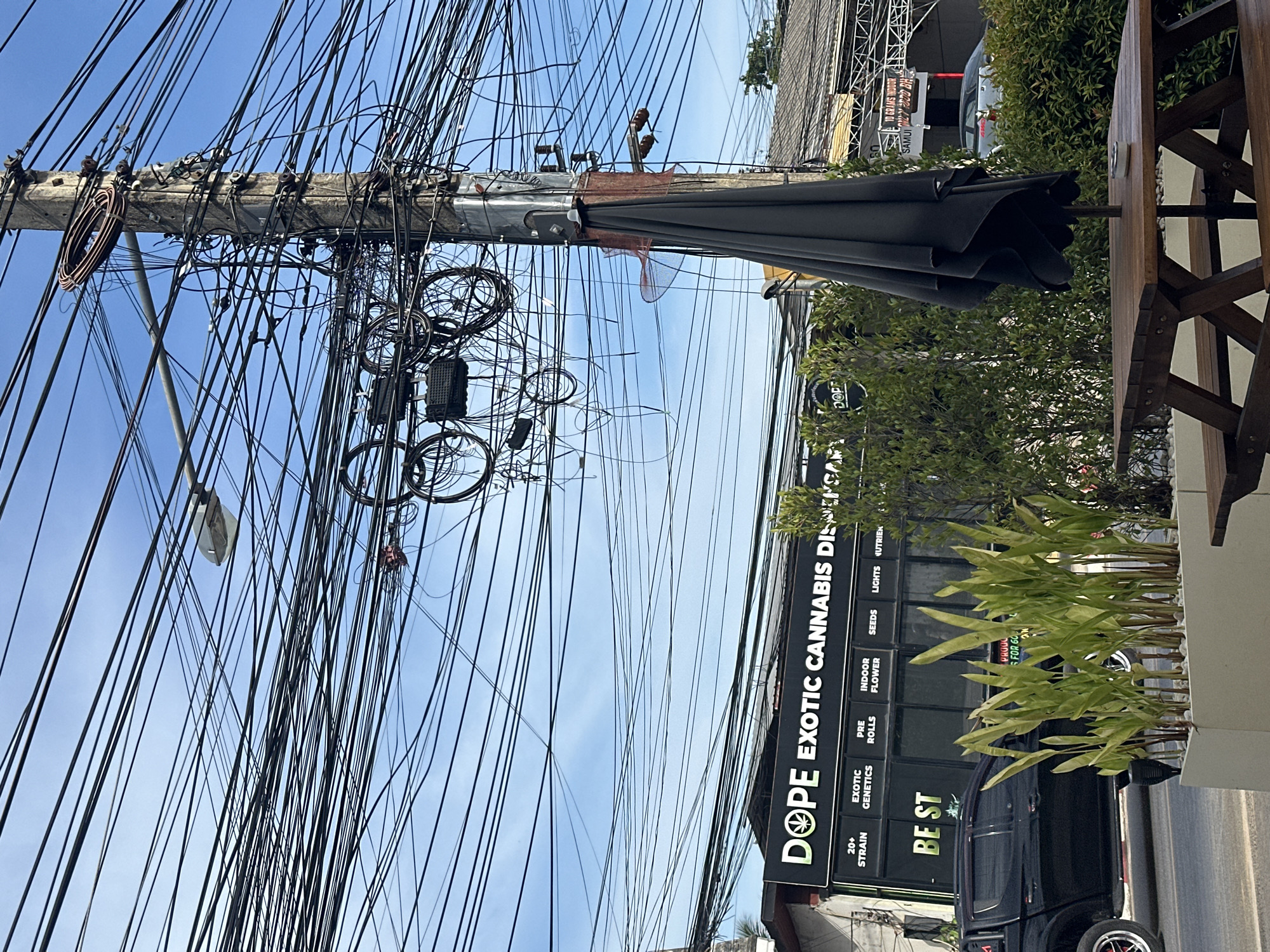 The image shows a street scene in the city of Ko Samui District, Thailand. A large utility pole with a chaotic tangle of black electrical wires and cables is prominent in the center, with a streetlamp attached to it. A dark grey umbrella is folded and tied to the utility pole. In the background, a building with a black facade and white lettering reads "DOPE EXOTIC CANNABIS DISPENSARY" is visible. Beneath the dispensary name, smaller white text indicates various products, including "20+ STRAIN," "EXOTIC GENETICS," "PRE ROLLS," "INDOOR FLOWER," "SEEDS," "LIGHTS," and "NUTRIENTS." A black pickup truck is parked to the left of the building. In the foreground, a white planter box contains several tall green plants. A wooden picnic table with benches is situated in front of the planter. The sky is blue with scattered clouds.