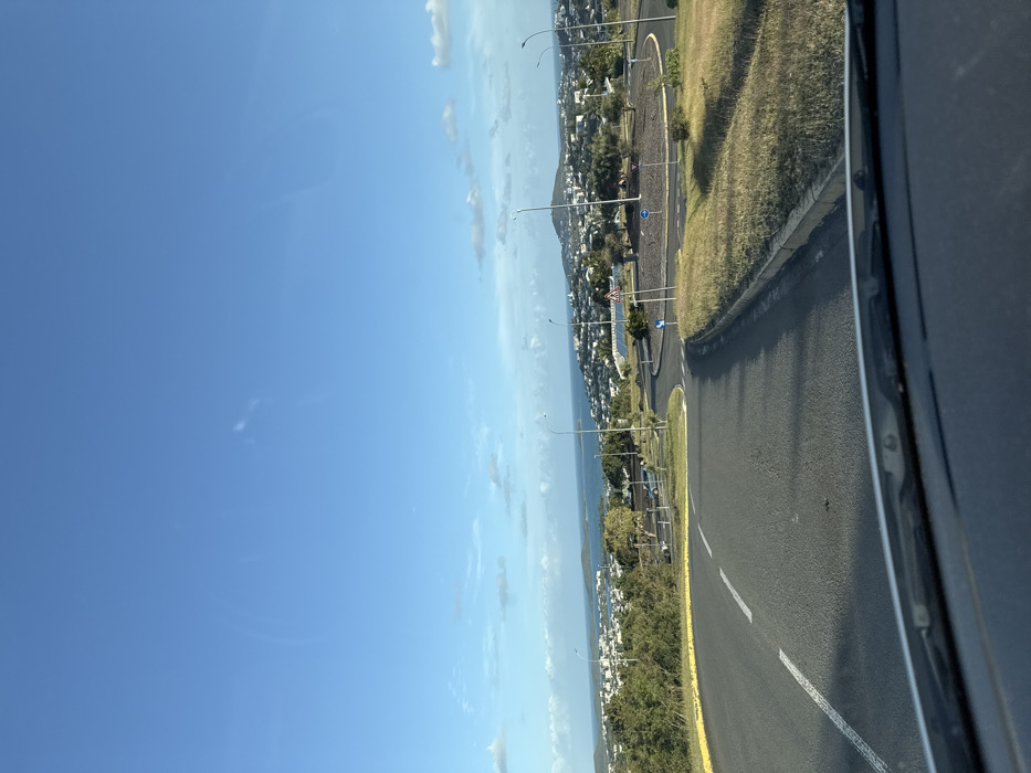The image depicts an elevated, daytime view from within a vehicle, with the top of the dashboard visible at the bottom of the frame, looking down a road towards a coastal city and bay in Nouméa, New Caledonia, an overseas territory of France.
The foreground features an asphalt road, marked with solid and dashed white lines, which descends and curves towards a multi-lane roundabout. To the left of the initial road, green foliage is visible. To the right, a strip of dry, yellow-brown grass separated by a concrete curb borders the road.
The midground is dominated by the circular roundabout, which has a central island covered with sparse, dry vegetation and several small trees. Multiple blue and white traffic signs, including a yield sign and a roundabout directional sign, are visible on the perimeter of the central island. Several streetlights with curved arms are positioned around the roundabout. Beyond the roundabout, to the left, additional road segments and a small parking area with parked vehicles are discernible.
The background reveals a sprawling urban landscape of predominantly light-colored buildings spread across green, undulating hills. Beyond the cityscape, a large, blue body of water, characteristic of a bay or ocean, stretches towards the horizon. Several landmasses or islands are visible within the water. The sky above is clear blue with scattered, wispy white clouds. No individuals or actively moving vehicles are present in the immediate foreground or midground.