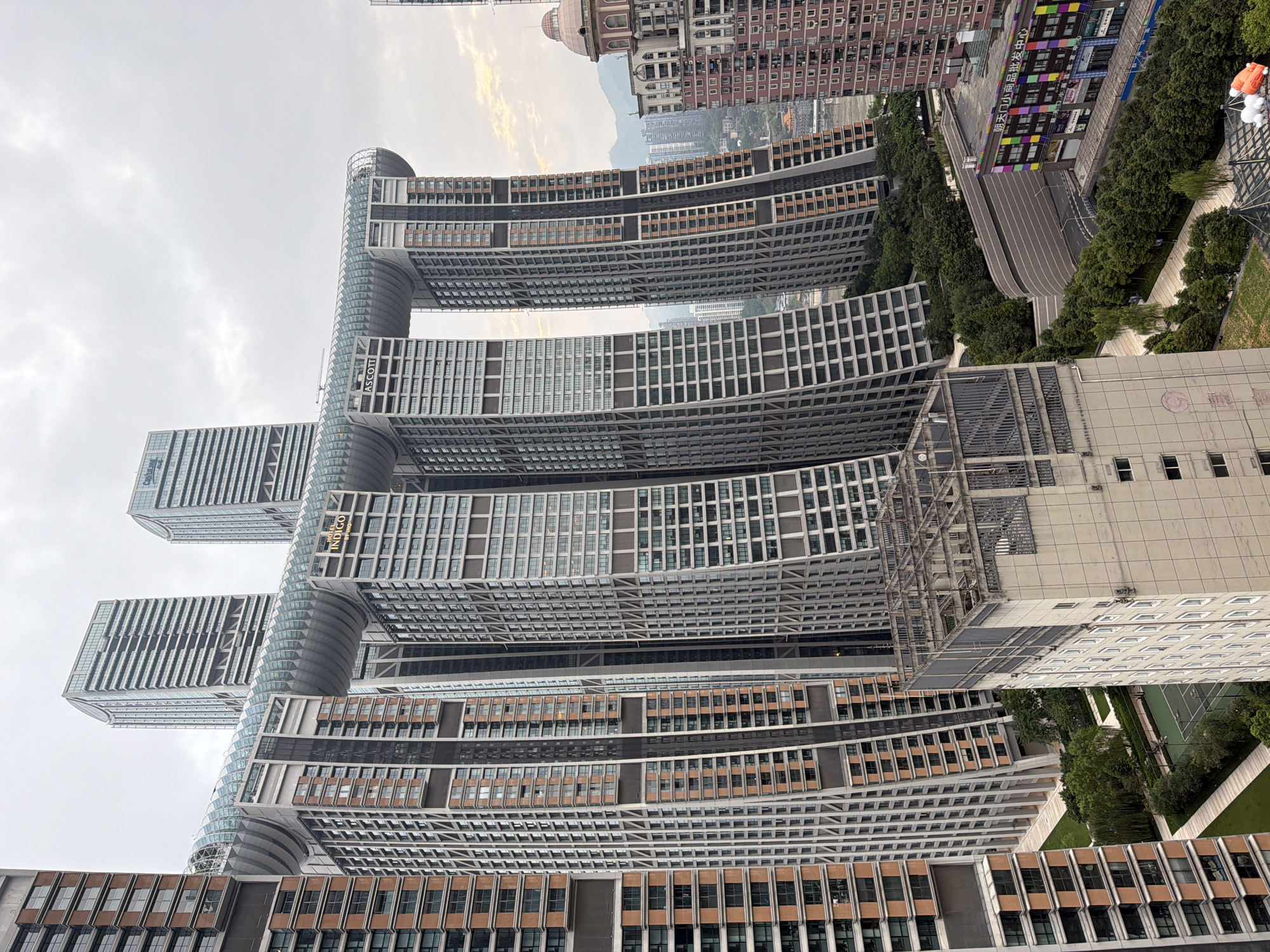 The image shows a close-up, eye-level aerial view of the Raffles City Chongqing complex, located in Yuzhong District, China. The complex is characterized by its striking architecture, featuring multiple skyscrapers connected by a horizontal skybridge. The skybridge is a prominent element, appearing as a long, undulating glass and steel structure that curves between the towers, resembling a ship or a dragon. The skyscrapers themselves are tall, slender buildings with predominantly glass facades, interspersed with sections of reddish-brown and grey panels, creating a modern, geometric aesthetic.
The foreground shows the upper sections of some of these towers, revealing their intricate window patterns and structural details. Below the skybridge, the buildings display a staggered, stepped design, giving the impression of immense scale. The skybridge's translucent covering allows glimpses of its internal structure and the sky beyond.
In the mid-ground, the tops of lush green trees and a neatly manicured park area are visible, including a tennis court. A white, multi-story building with a concrete-like facade and a series of windows occupies the lower right portion of the image. Further into the background, other buildings of varying heights and architectural styles are scattered across the urban landscape, with distant hills or mountains visible under a cloudy sky. A brightly colored sign with Chinese characters is visible on one of the lower buildings, adding a touch of vibrancy to the scene. The overall atmosphere is one of urban grandeur and modern architectural innovation.