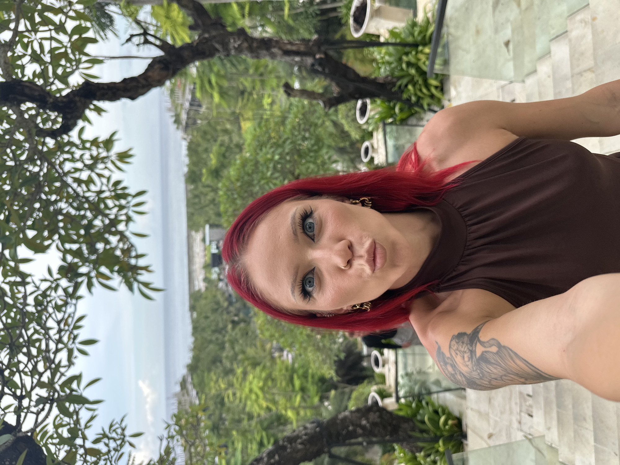 A woman with red hair, fair skin, and blue eyes is taking a selfie. She is wearing a brown top. Her right arm is visible, showing a tattoo of a bird or angel on her forearm. The background includes lush greenery, trees, and a view of the ocean. The setting appears to be a balcony or elevated area with glass railings and stone steps. The city is Nusa Dua, Indonesia.