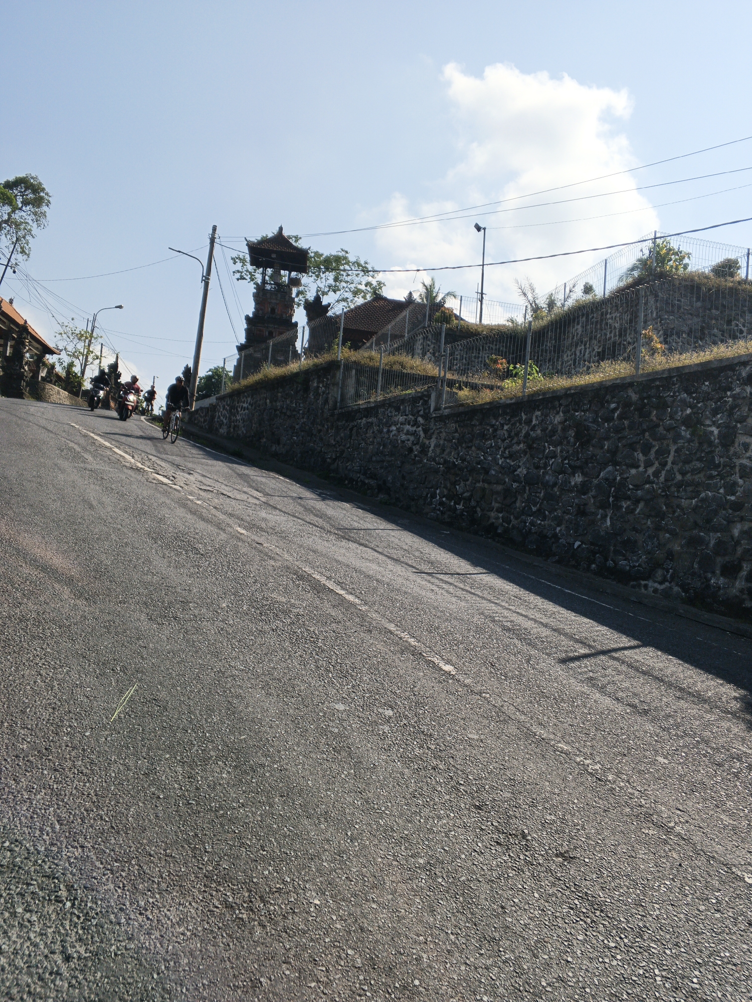 In Abiansemal, Indonesia, a paved, uphill road is depicted under a bright, partially cloudy sky. A man on a bicycle is riding up the incline, followed by three individuals on motorcycles. On the right side of the road, a stone wall supports a fenced grassy embankment. Above the embankment, a traditional Balinese structure with a multi-tiered roof is visible, surrounded by trees and utility poles with electrical wires. Buildings with red tiled roofs are also present on the left side of the road, some with stone accents. The road surface is asphalt with a white dashed line marking its center.