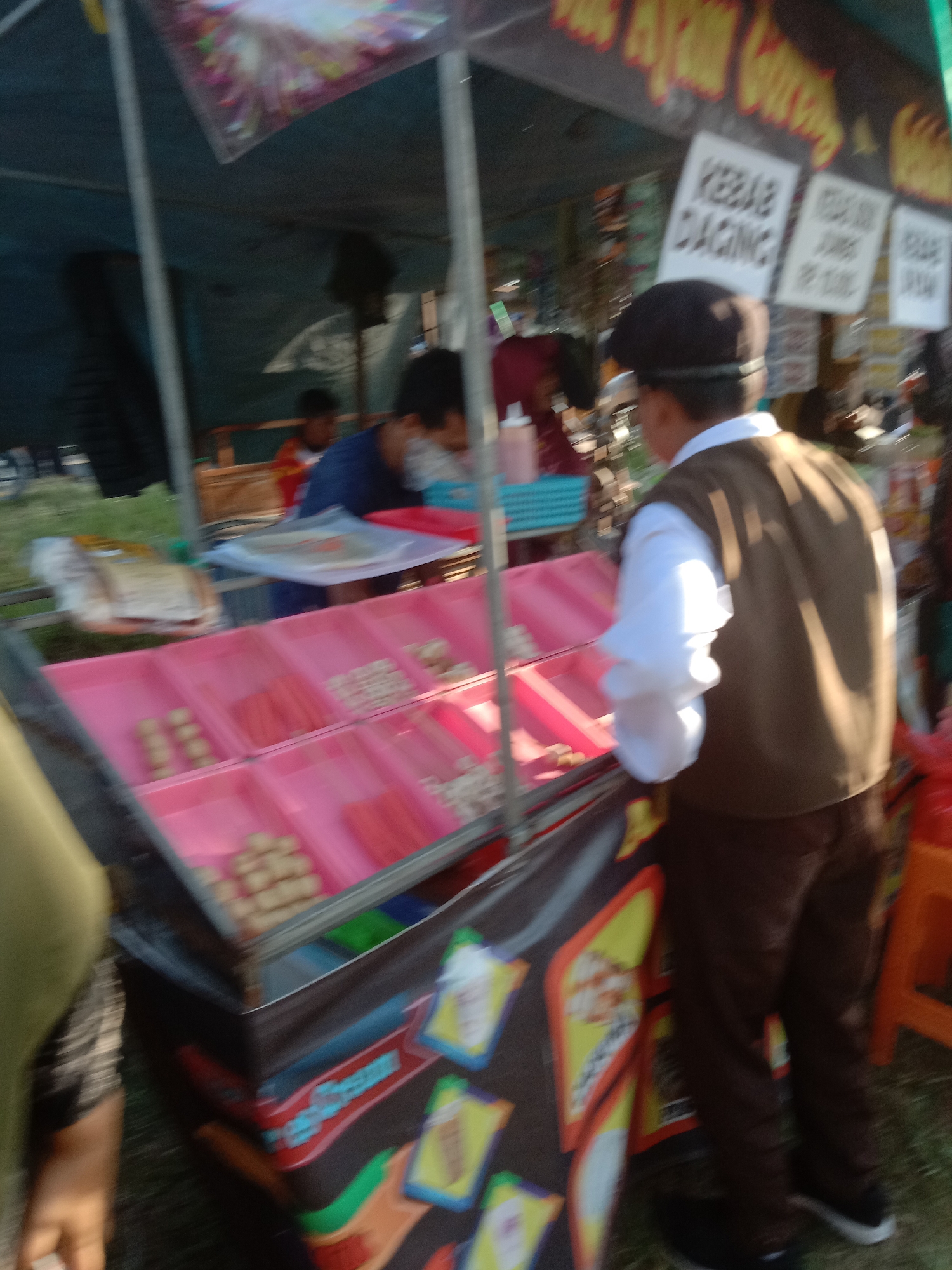 A vendor, wearing a brown vest over a white shirt and a dark cap, stands behind a food cart in what appears to be an outdoor market. The cart has pink display trays filled with various food items, including what look like meatballs and processed meat sticks. Signs with Indonesian text, including "KEBAB DAGING," are visible on the cart. In the background, other people are present, and the general environment suggests a bustling marketplace. The image is blurry, making precise identification of all details difficult.