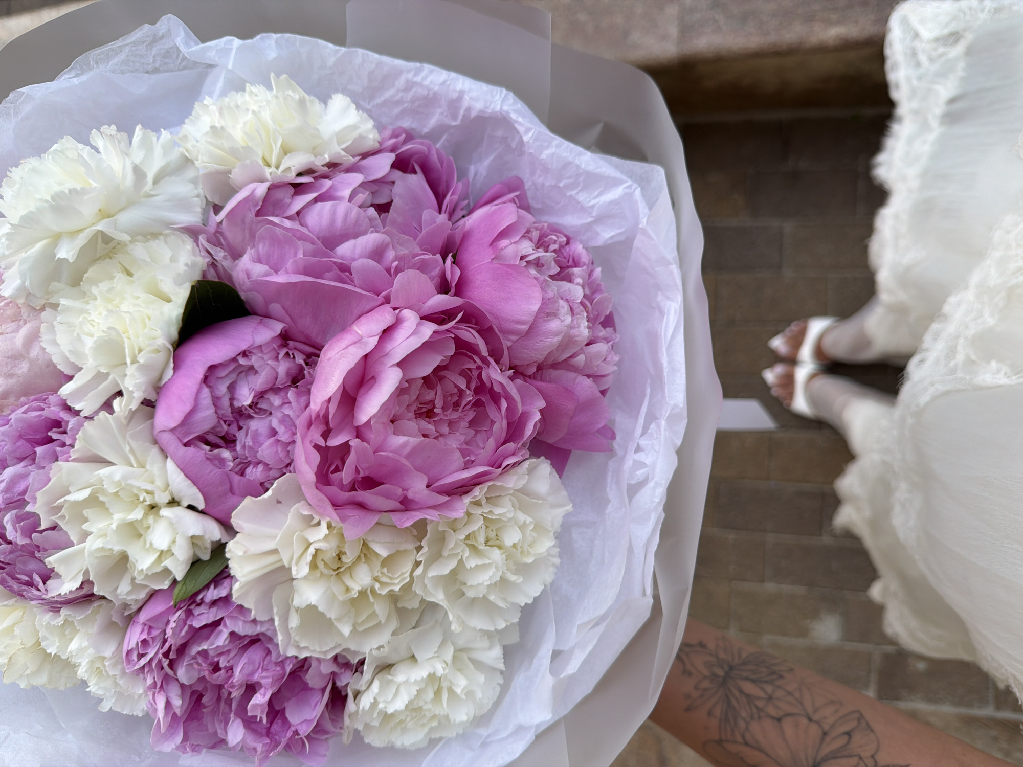 A close-up, high-angle shot captures a bouquet of pink peonies and white carnations, artfully arranged and wrapped in white tissue paper. The flowers are the primary focus, with their delicate petals and vibrant colors filling most of the frame. To the right of the bouquet, a person's legs are visible, adorned in a white, ruffled gown and white heeled sandals. Their feet are positioned on a tiled floor, suggesting an outdoor setting. A subtle tattoo of floral designs is visible on the person's lower leg. The overall impression is one of elegance and celebration, possibly a wedding or special occasion. The lighting suggests it could be daytime. There is no visible text in the image. The location is Novaya Usman, Russia.