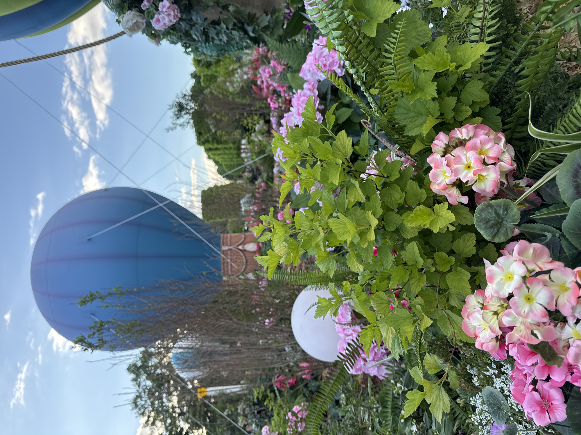 The image shows a blue hot air balloon floating in the sky in Moscow, Russia. The balloon is partially obscured by greenery and flowers, including pink and white blossoms, ferns, and leafy plants. A white spherical object is also visible amidst the vegetation. The sky is blue with some clouds.