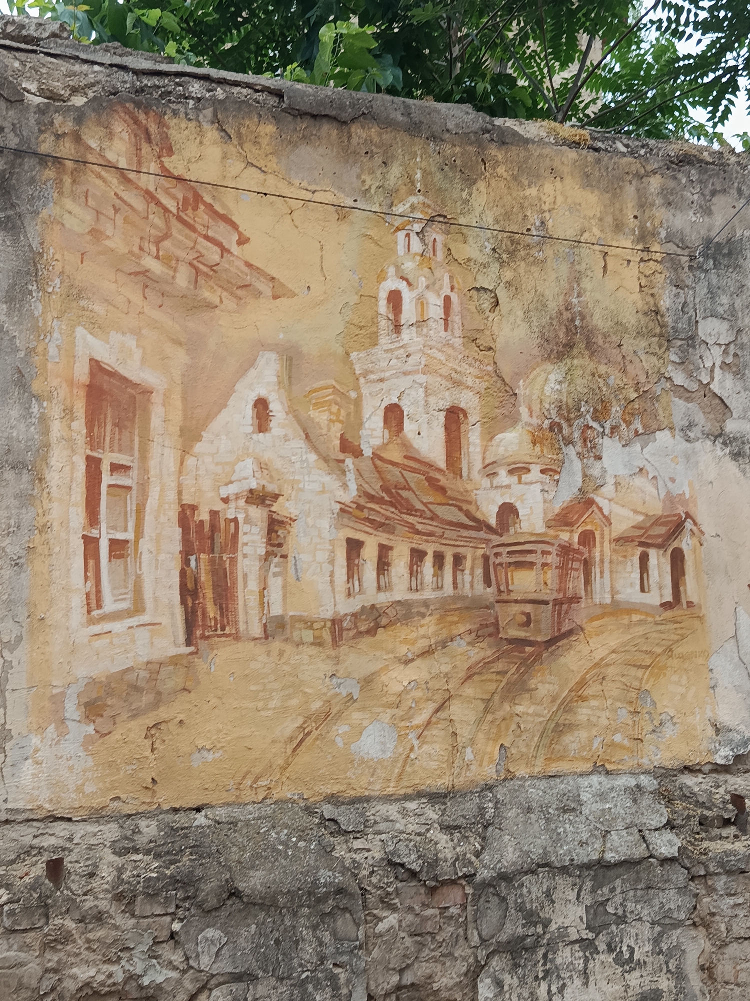 A mural depicting a European-style cityscape adorns a crumbling concrete wall. The artwork, rendered in muted tones of ochre, white, and brown, features buildings with arched windows and doorways, a prominent church with a bell tower, and what appears to be a tram or trolley car on tracks. The paint is peeling and faded in many areas, indicating age and exposure to the elements. The wall itself is textured and shows signs of significant decay, with cracks and missing sections of plaster. At the top of the image, green foliage from trees overhangs the wall. Below the mural, a lower section of the wall is constructed from rough, aged bricks and mortar, further emphasizing the dilapidated state of the structure. The scene is static and conveys a sense of history and abandonment.