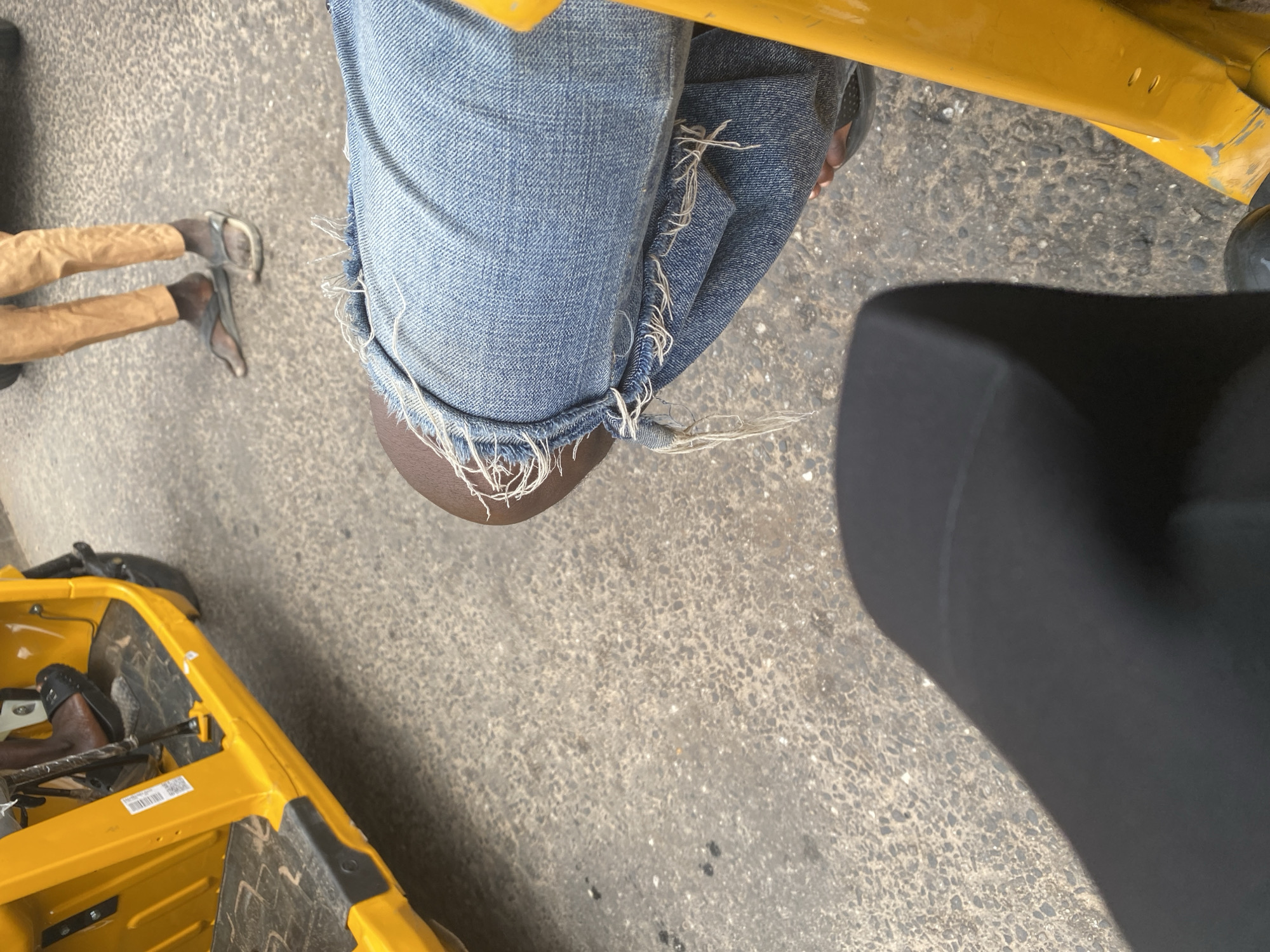 The image shows a person's legs and torso from a high-angle perspective, with a yellow object and a black object in the foreground. The person is wearing blue jeans with frayed edges and tan pants. Their feet are clad in dark sandals. They appear to be sitting in or on a yellow vehicle, possibly a tuk-tuk or auto-rickshaw, which is partially visible at the bottom left of the frame. The vehicle's interior appears to be dark. A black, cushioned object, likely a seat or part of the vehicle, is prominent on the right side of the image, casting a shadow.
The ground is a textured, grey asphalt surface with scattered small white and dark specks, suggesting an outdoor environment. The scene is well-lit, indicating daytime, likely sunny given the distinct shadow cast by the black object. The overall impression is one of casual daily life. The frayed edges of the jeans and the simple footwear suggest an informal setting. No specific emotions are discernible from the image.
There is a small white sticker with black text on the yellow vehicle, but the text is illegible. The location context provided is Jalingo, Nigeria.