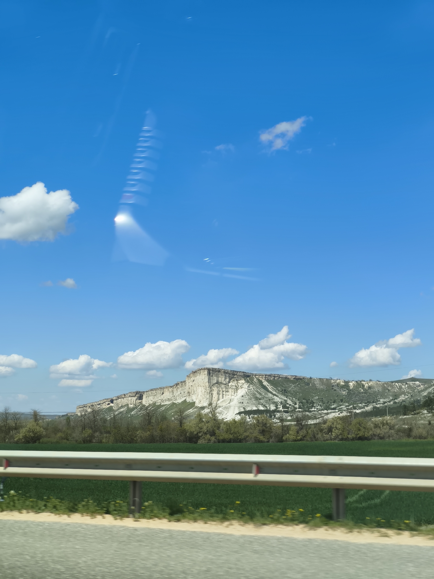 The image shows a landscape under a bright blue sky with scattered white clouds. In the foreground, a silver guardrail runs parallel to the bottom of the frame, separating a green field with yellow wildflowers from the road. Behind the field, a large white rock formation rises from the ground, covered with green vegetation on its upper slopes. The image is taken from a moving vehicle. The city is Unknown, Unknown.