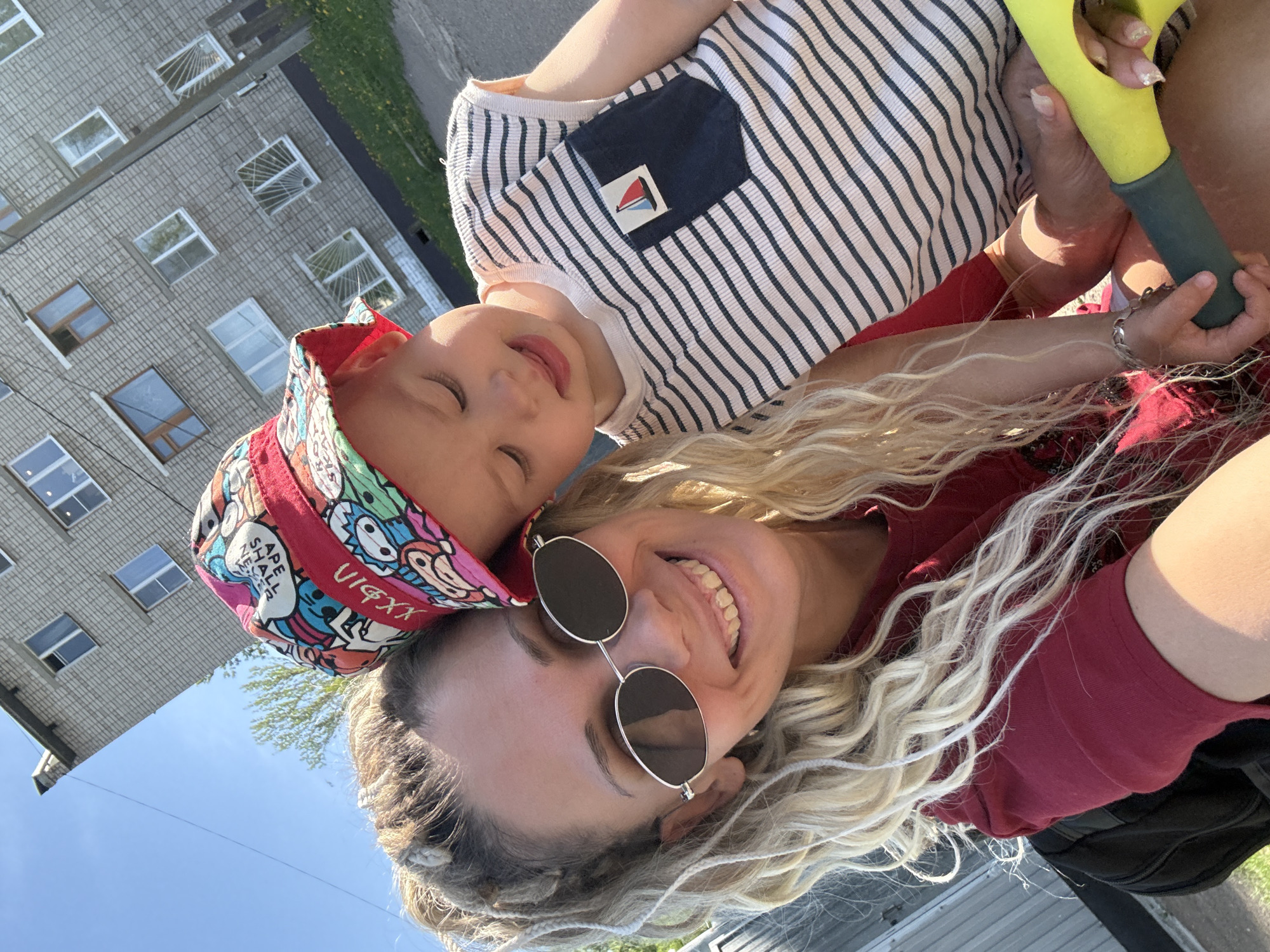 A young woman with long blonde hair, wearing round sunglasses and a red shirt, is holding a young child with a colorful patterned hat and a striped shirt. They appear to be outdoors, possibly in an urban area, with a building visible in the background. The child's eyes are closed as the woman smiles broadly. She is holding a yellow and gray object, likely a child's toy.