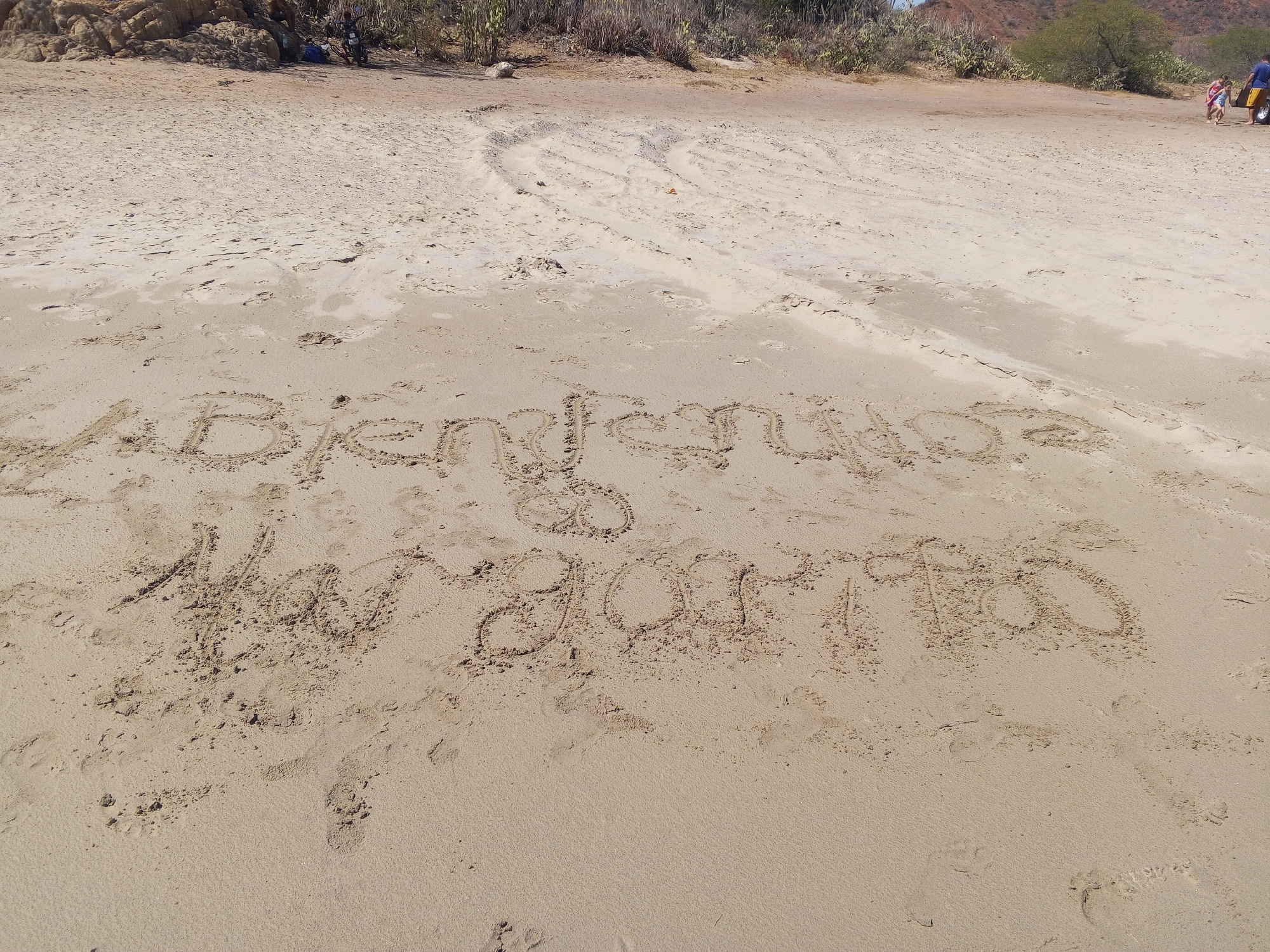 Scene: A sunny beach in Juan Griego, Venezuela.
Environment: Sandy beach, light-colored sand, some vegetation at the back, slight incline in the terrain.
Text: The text "ABkendiciones Margarita 9-08-1980" is written in the sand.
Objects: Footprints and tire tracks are visible in the sand.
People: Two individuals and what appear to be a yellow cart are present in the background, near the trees.