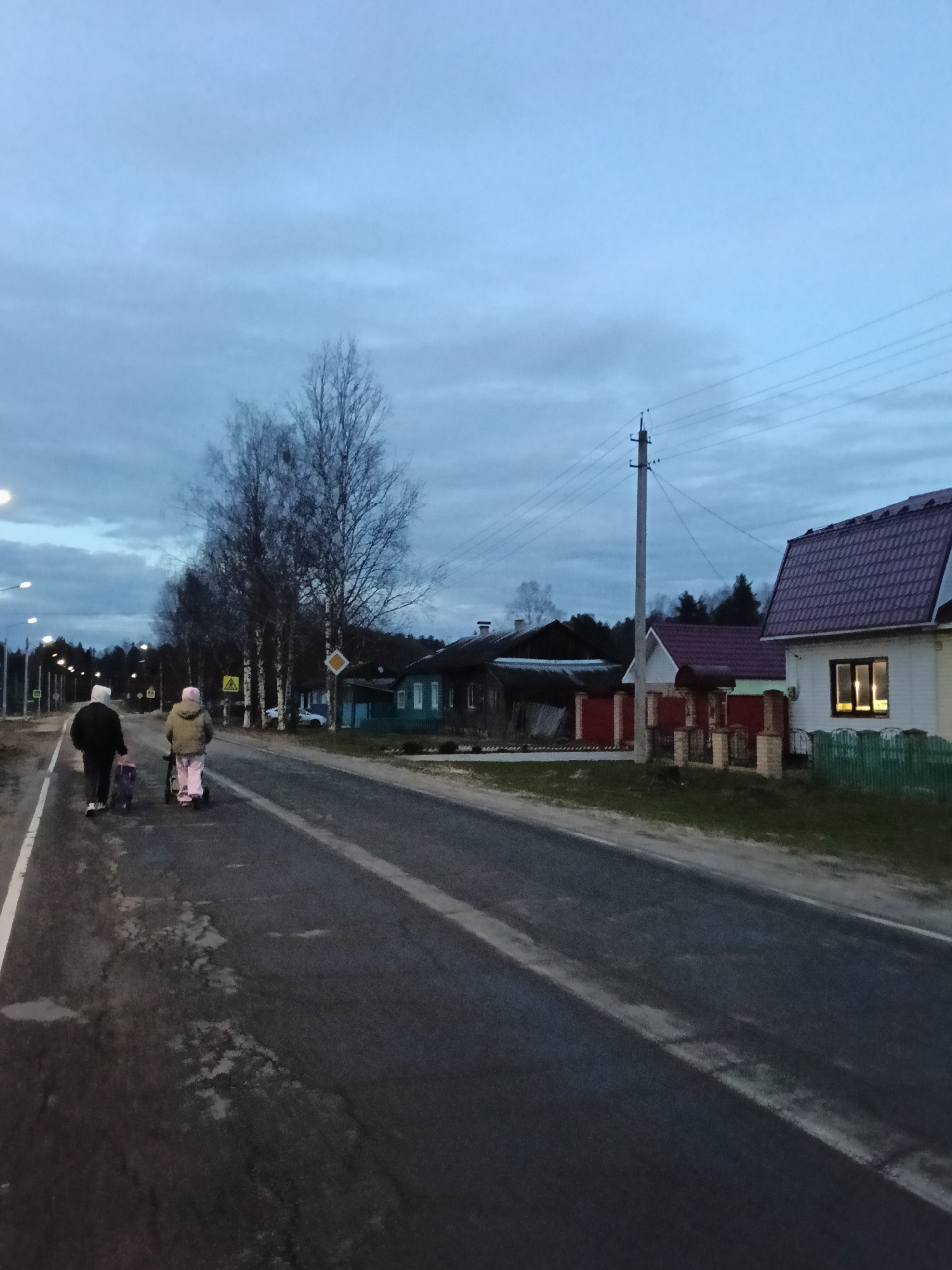 Two individuals are walking along a road in Красный Маяк, Russia. The person on the left is wearing a dark coat and carrying a bag, while the person on the right is wearing a light-colored jacket and pushing a stroller. The road has a white line marking the edge, and the pavement is cracked. On the right side of the road are several houses, some with visible lights in the windows. A utility pole stands near the houses. The sky is overcast, suggesting it is either early morning or late evening. Bare trees stand between the road and the houses. A yellow road sign is visible near the trees.