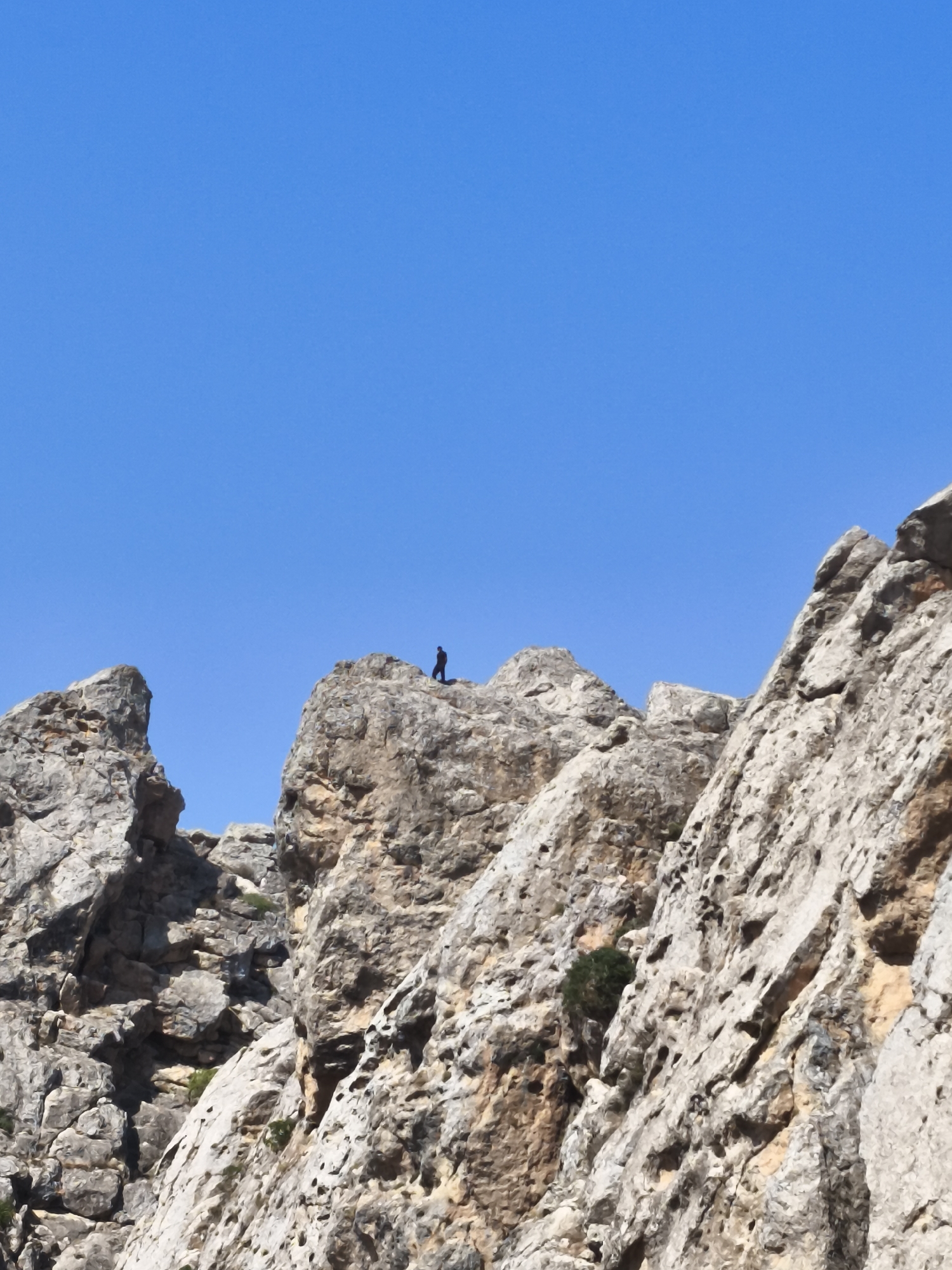 A single, silhouetted figure stands on a rocky outcrop against a clear blue sky. The figure, appearing small in comparison to the imposing rock formations, is facing away from the viewer. The foreground is dominated by rugged, textured rocks, indicative of a mountainous or canyon-like environment. Hints of sparse vegetation can be seen clinging to the rocks. This scene is located in Qalaşıxı, Azerbaijan, a region known for its dramatic geological landscapes.