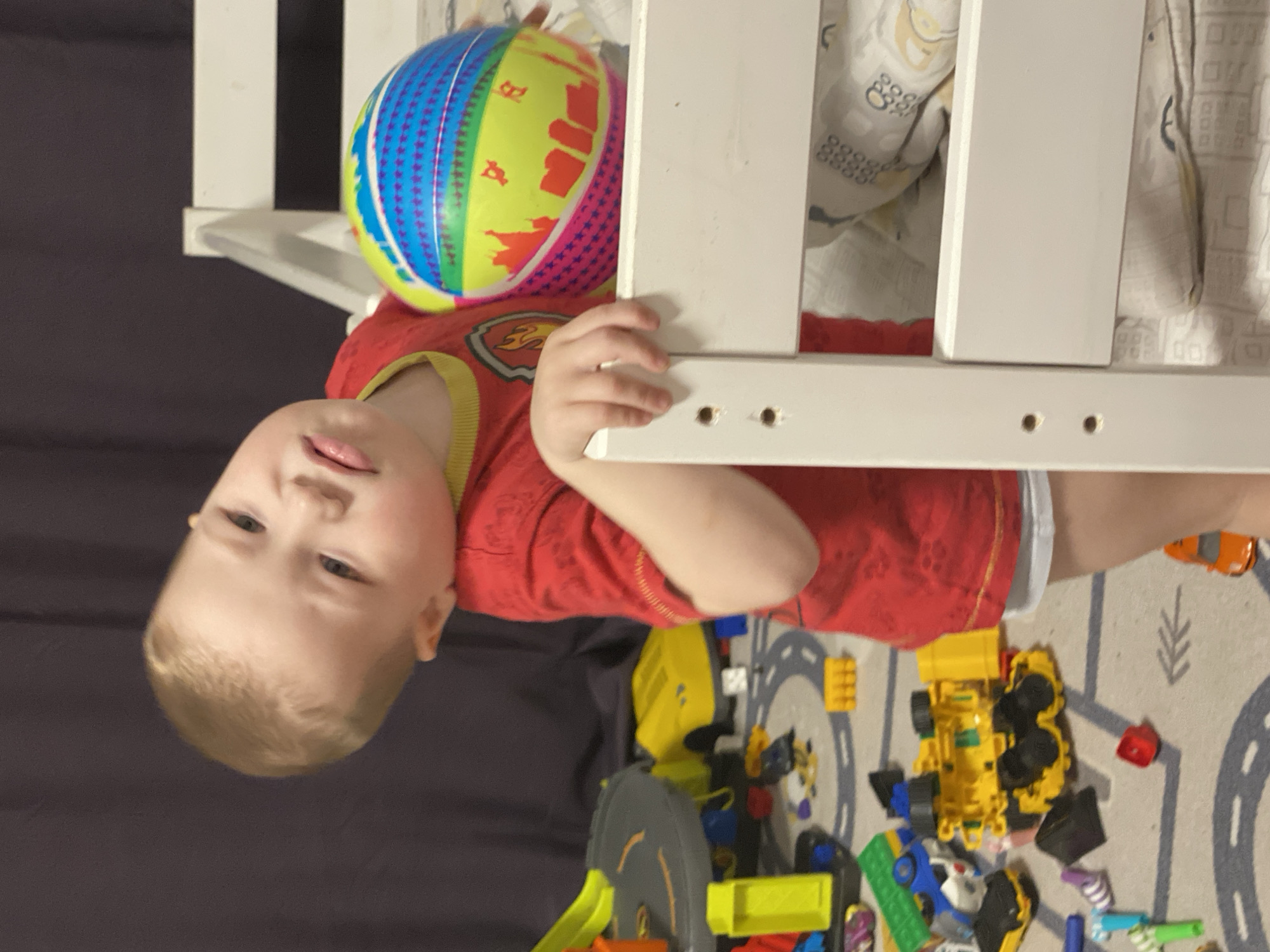 The image depicts a young child indoors, likely within a bedroom or play area. The specific location within the city of Sochi, Russia, cannot be determined from the visual information provided.
The central subject is a fair-skinned child, approximately two to three years old, with short, light brown or blonde hair. The child is wearing a red short-sleeved t-shirt with a yellow collar and a "Paw Patrol" emblem on the left chest, and a white diaper or underwear visible below the shirt. The child's head is tilted to the left, and they are looking directly forward with a neutral or slightly pouting expression. The child's right hand rests on a white wooden bed frame, which appears to be part of a bunk bed or crib. In the child's left arm, they hold a colorful, inflatable beach ball featuring wide horizontal stripes in yellow, green, blue, pink, orange, and purple.
The background shows a cluttered play area. The floor is covered with a light-colored rug featuring a grey road map pattern. Numerous children's toys are scattered across the rug, including a grey and yellow multi-level toy garage or ramp, several yellow toy vehicles (such as a bulldozer or dump truck), various plastic blocks in colors like blue, green, red, and black, and other small plastic play items. A dark grey or black curtain or fabric backdrop is visible in the upper left background. The white wooden bed frame continues to the right, revealing a mattress and a patterned sheet (white with yellow and grey motifs) on the lower bunk or crib. The scene is illuminated by ambient indoor lighting.