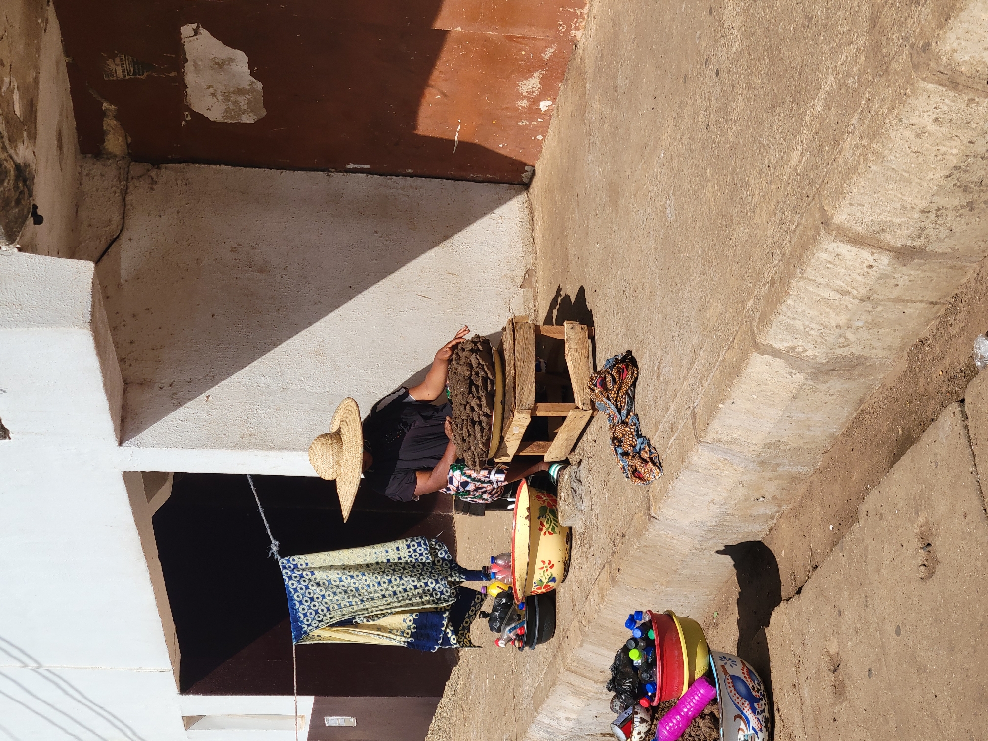 The image captures an overhead view of an outdoor scene, likely an alleyway or a small courtyard in Bukuru, Nigeria, bathed in bright sunlight. The setting appears to be a functional, possibly informal, workspace or market stall area.
The primary subject is a person, seen from behind, seated on a low wooden stool or crate. They wear dark clothing, and a straw hat rests near them, possibly on their head or an adjacent surface. Next to the person is a large, irregularly shaped pile of dark brown, organic-looking material, suggesting they are engaged in sorting, processing, or preparing something.
Surrounding the person are various objects related to what could be a vending or recycling operation. A patterned blue and white fabric is draped over a rope strung across the space, potentially for display or drying. Another colorful fabric, with orange, brown, and blue patterns, is laid on the ground near the wooden structure. Several bowls are visible: a large, yellow enamel bowl with floral decorations, and smaller, brightly colored plastic bowls (red, yellow) filled with an assortment of plastic bottles and loose plastic caps, indicating a collection of recyclables or items for sale.
The ground is an uneven mix of dirt and possibly rough concrete. The surrounding walls are made of concrete or stucco, with sections painted white and others a deep reddish-brown. There are visible cracks, peeling paint, and general wear on the wall surfaces. A small, faded piece of paper with unreadable text is barely discernible on one of the brown walls. Strong, sharp shadows cast by the buildings and objects indicate a sunny day, likely mid-morning to early afternoon.