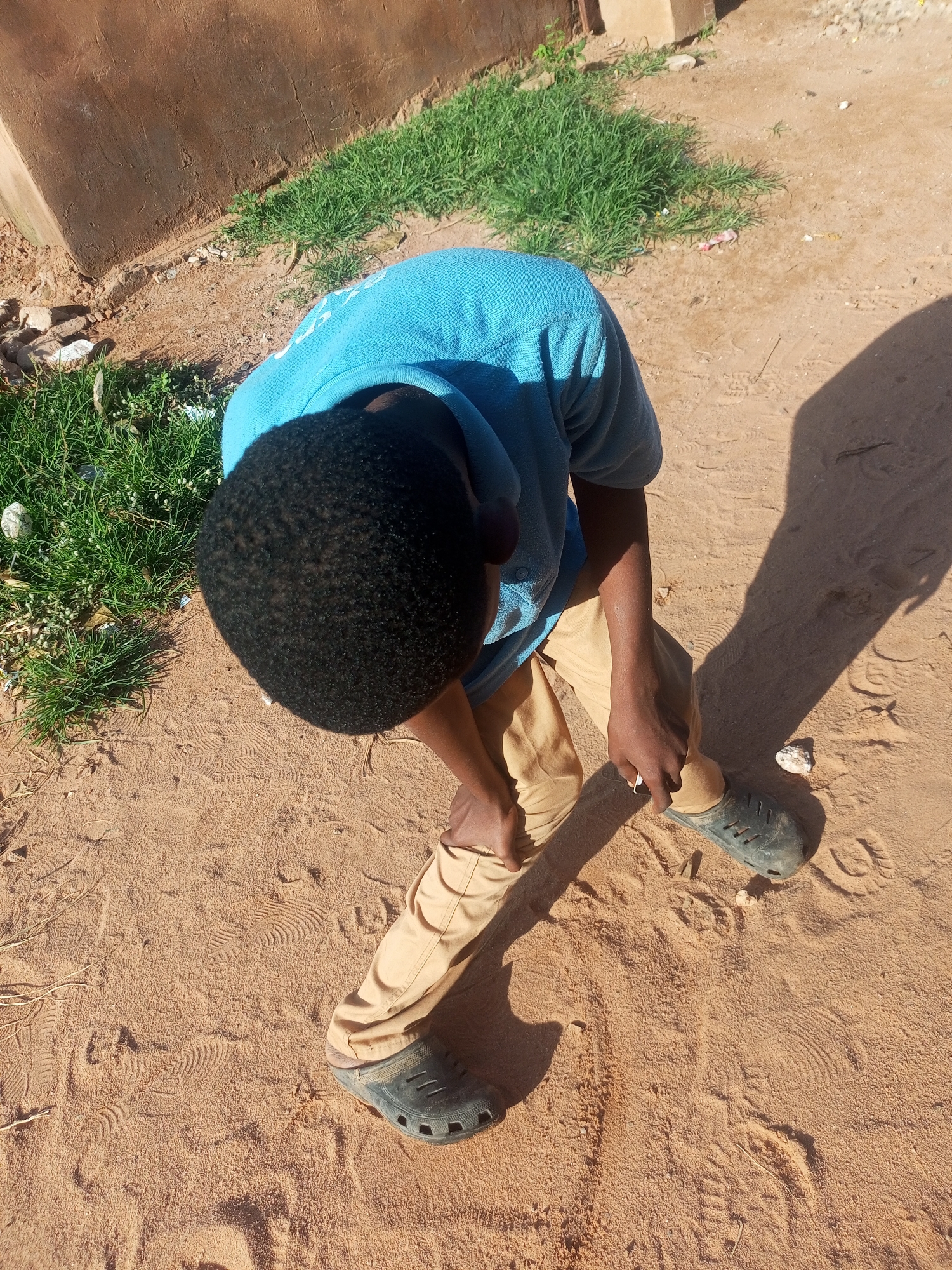 A young boy, with dark skin and short black hair, is kneeling on a sandy ground in what appears to be the city of Tempure, Nigeria. He is wearing a blue t-shirt and tan pants, along with black slip-on shoes. The boy is looking down and appears to be adjusting his shoe. To the left of the boy, there is a patch of green grass growing next to a brown wall. The ground is mostly sand, with some footprints visible. The lighting suggests it is daytime, with a distinct shadow cast by the boy.