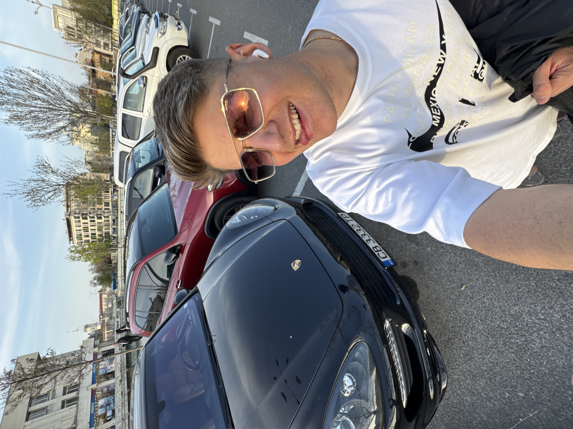 A man takes a selfie outdoors next to a black Porsche Cayenne. The car’s license plate is visible. Other cars of various colors and models are parked nearby. The man is wearing sunglasses, a white t-shirt with text, and an earbud. The image was taken in the city of Plovdiv, Bulgaria. Buildings are in the background, and the sky is visible.
