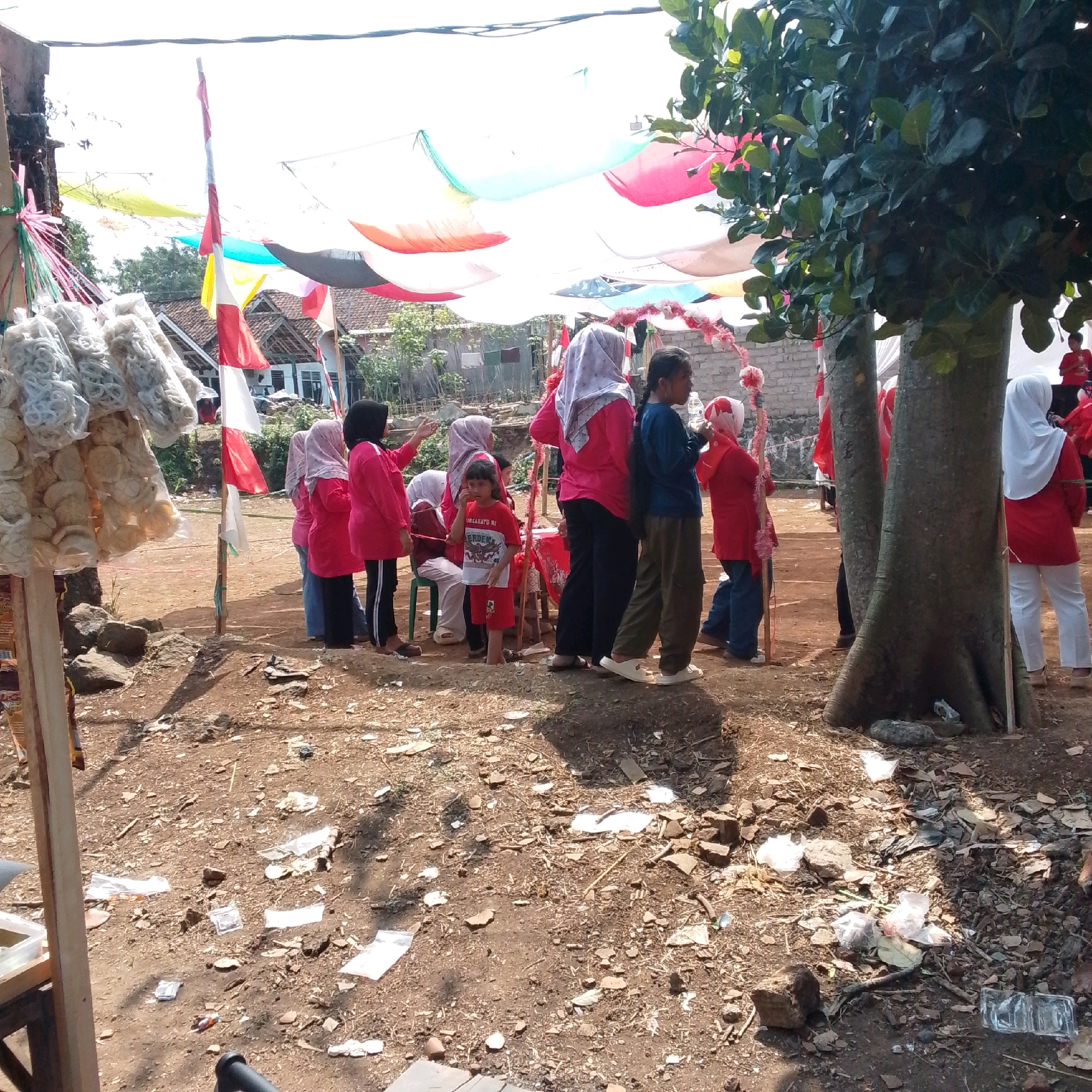 The image depicts an outdoor scene in Cilaku, Indonesia, characterized by a gathering of people, possibly for an event. A group of individuals, predominantly women and children, are standing and interacting in a dusty, outdoor area. Some are wearing colorful clothing, including headscarves. In the foreground, bags of what appear to be crackers or snacks are displayed on a makeshift stand, indicating a possible vendor or food stall. Indonesian flags, red and white, are visible, suggesting a national celebration or event. Overhead, fabric sheets are strung up, creating a canopy effect, perhaps for shade or decoration. The environment appears somewhat rustic, with dirt ground littered with debris. The background reveals residential structures and greenery. The overall impression is one of a local community event taking place on a sunny day.