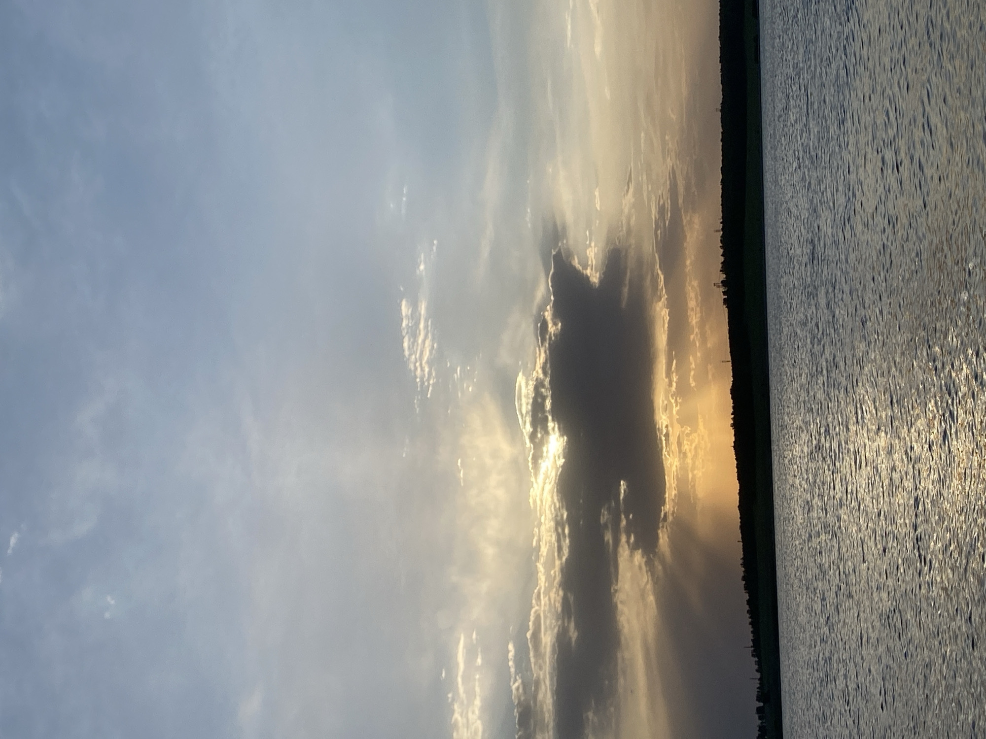 The image depicts a view of the lake and sky in Smolino, Russia. The sky is mostly blue with a large cloud formation obscuring the setting sun, creating sunrays. The landscape is a strip of trees, and the lake water is rippled with light reflecting on the surface.