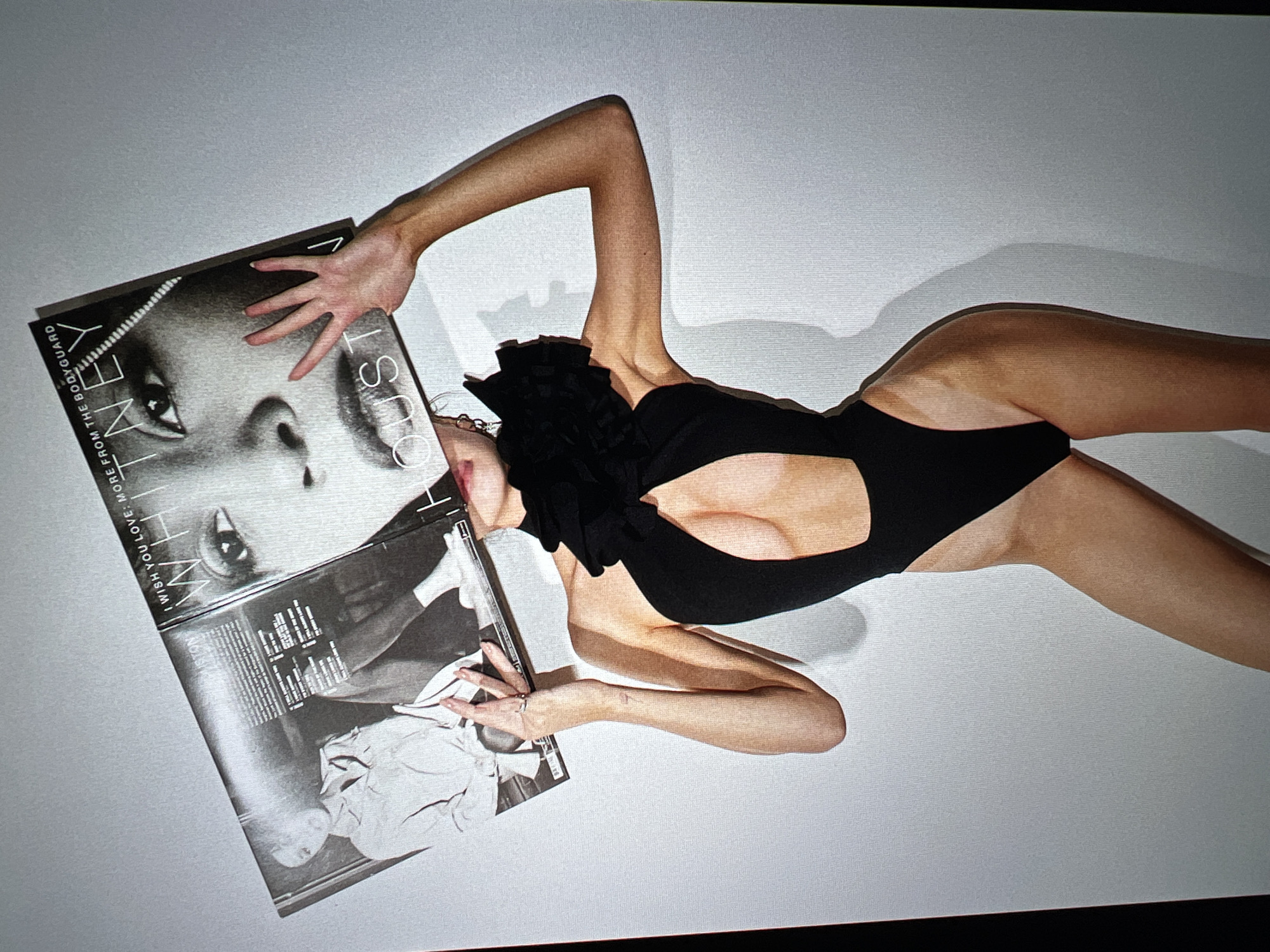 A person is holding a Whitney Houston album in front of their face against a white background. The person is wearing a black swimsuit with a cutout and ruffle detail. The album cover features a close-up of Whitney Houston's face. The person's hands are visible on the album.