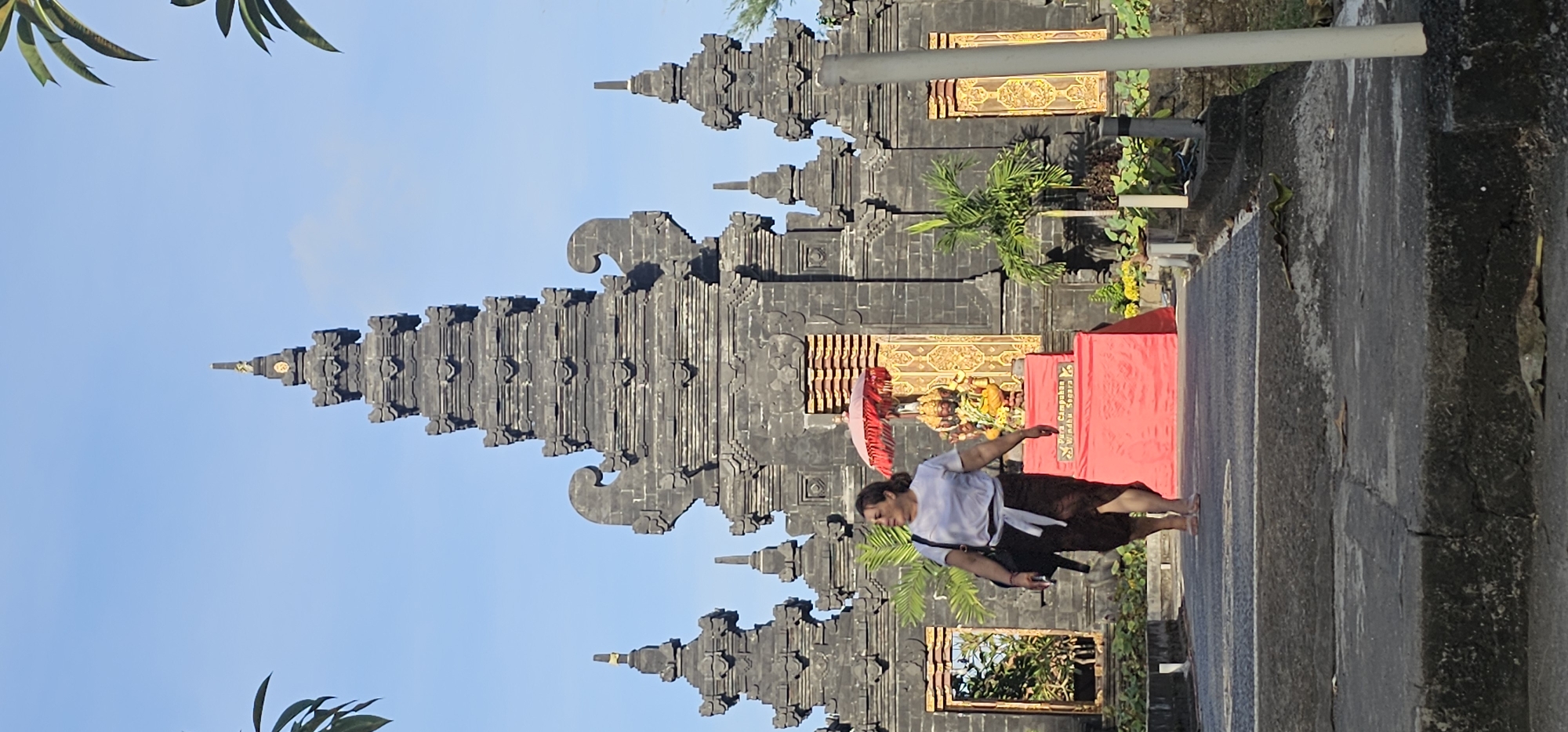 A full shot depicts a person performing a dance in front of a traditional Balinese structure, likely a temple gateway, on a sunny day. The person is dressed in a white top, a dark brown sarong, and a white sash around their waist. They are barefoot and appear to be mid-movement, with one arm extended to the side. Behind them is an ornate, gray-stone Balinese gateway, with multiple levels of detailed carvings and spires. A small altar with a red cloth, offerings, and a red umbrella sits in front of the gateway. The altar is adorned with decorations and flowers. Lush greenery and other plants surround the gateway, suggesting a tropical location. The sky is a clear blue with a few scattered clouds. Based on the traditional Balinese architecture, it's highly probable that the location is in or near Denpasar, Indonesia.