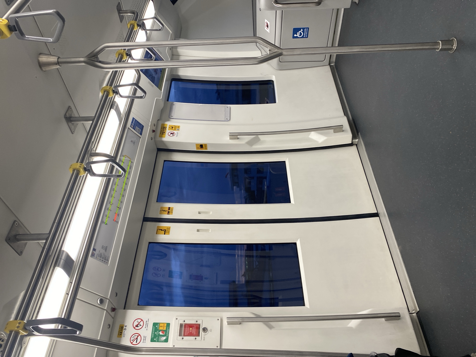 The image shows the interior of a train car in Bang Mueang Subdistrict Municipality, Thailand. The scene is focused on the train doors, which are white with blue-tinted windows. There are safety signs and emergency controls near the base of the doors. Handrails and overhead bars are visible, suggesting a public transportation setting. The floor is gray, and accessibility symbols are present, indicating accommodations for disabled passengers. No people are visible in the shot.