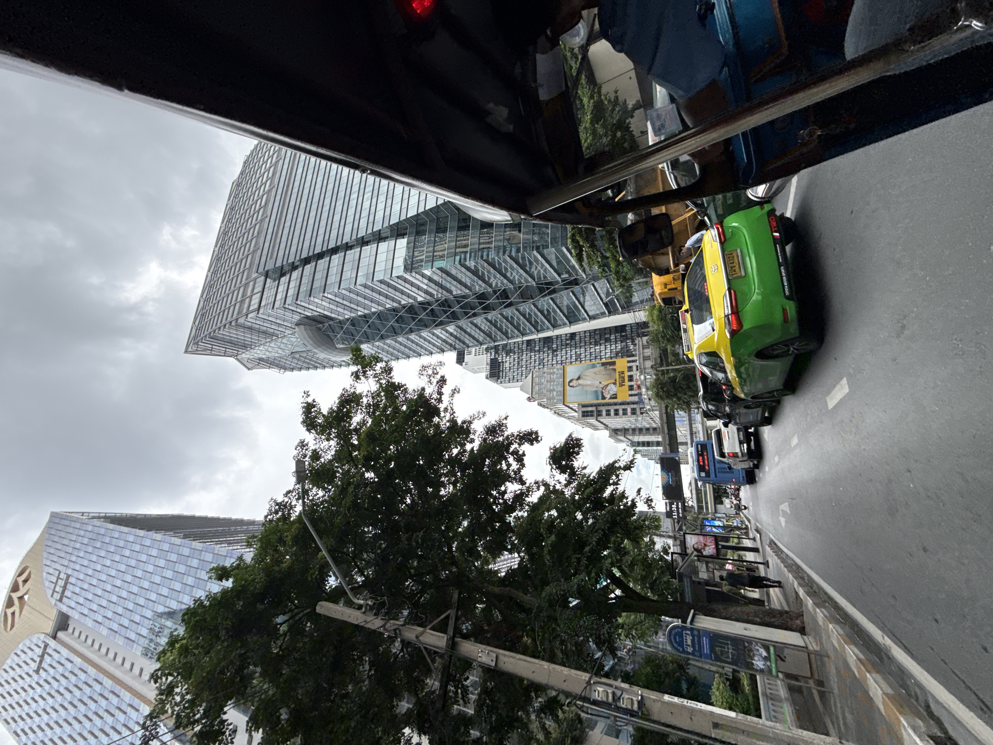 The image depicts a street scene in Bangkok, Thailand, likely in a business district due to the presence of modern skyscrapers. The perspective is from a vehicle, possibly a tuk-tuk, looking forward and slightly to the right. The sky is overcast.
In the foreground, the right side of the image is dominated by the interior of a vehicle, with dark framing and a hint of blue upholstery. A red light is visible on the top right.
The street itself is wet, suggesting recent rain. Traffic is present, with several vehicles visible. A green and yellow taxi is prominent in the center, its rear end facing the camera. A white pickup truck is in front of it, and further ahead, a blue bus. To the left of the road, there is a sidewalk with a bus stop shelter and a utility pole adorned with wires and a street lamp. Trees with dense foliage line the sidewalk, obscuring parts of the buildings behind them.
The background features several tall, modern buildings with glass facades. One building on the left has a distinctive curved top with decorative elements. Another building on the right has a striking, angular design. A large billboard with an image of a woman and text is visible on one of the buildings.
On the sidewalk, a person in dark clothing is walking away from the camera. Another person, wearing a light blue shirt, is visible near the yellow taxi, seemingly looking at something on the ground or the vehicle.