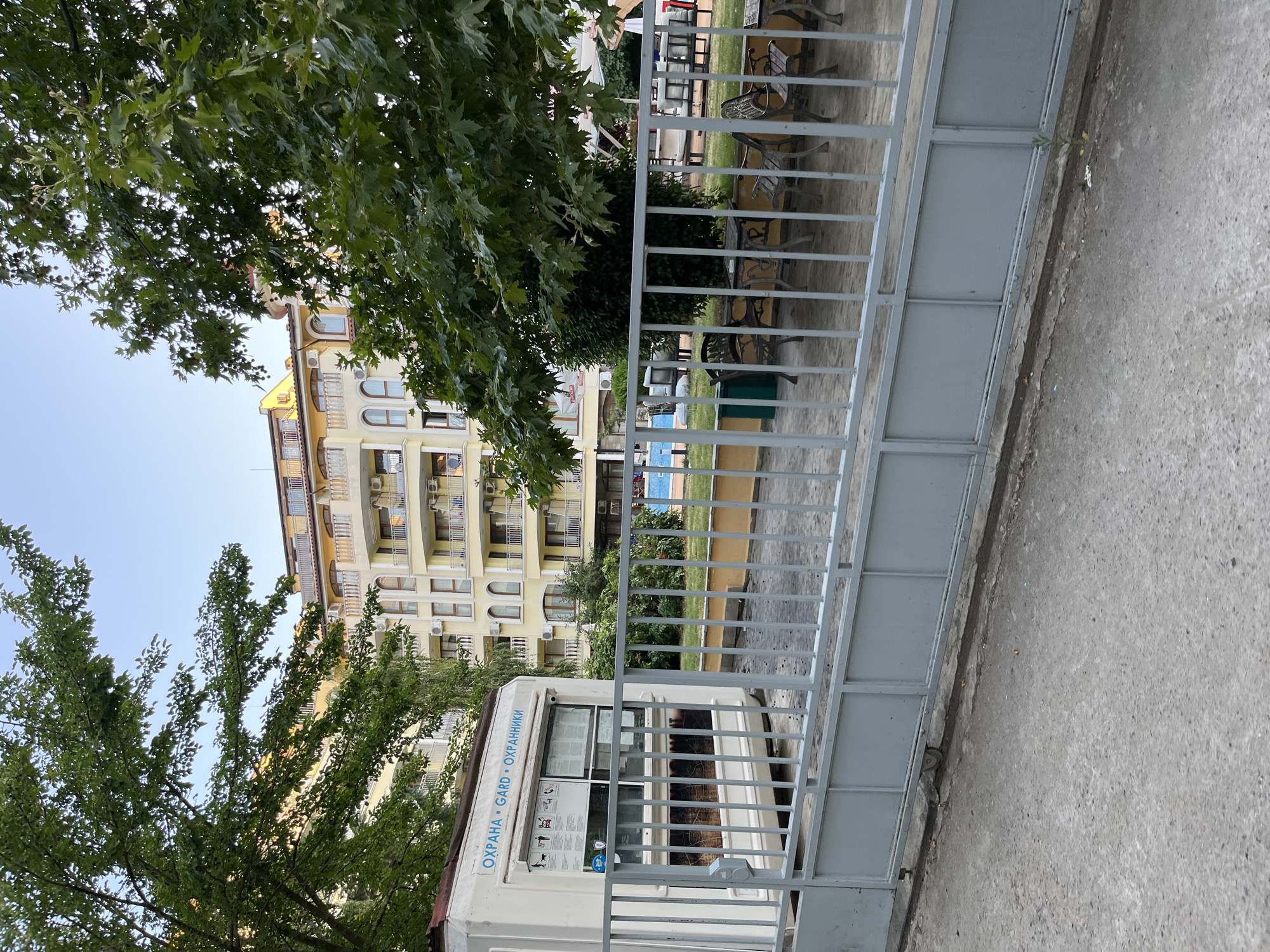 The image shows a gated entrance to a residential complex in Nesebar, Bulgaria. A white security booth with the words "ОХРАНА • GARD • ОХРАННИКИ" (meaning "Protection • Guard • Guards" in Bulgarian and English) is visible on the left. Beyond the gate, a yellow apartment building with balconies and arched windows stands tall. A swimming pool and outdoor seating areas with tables and chairs are set up in the courtyard, surrounded by lush green trees and foliage. The sky is clear and blue, suggesting a pleasant day. The overall scene depicts a tranquil and well-maintained property.