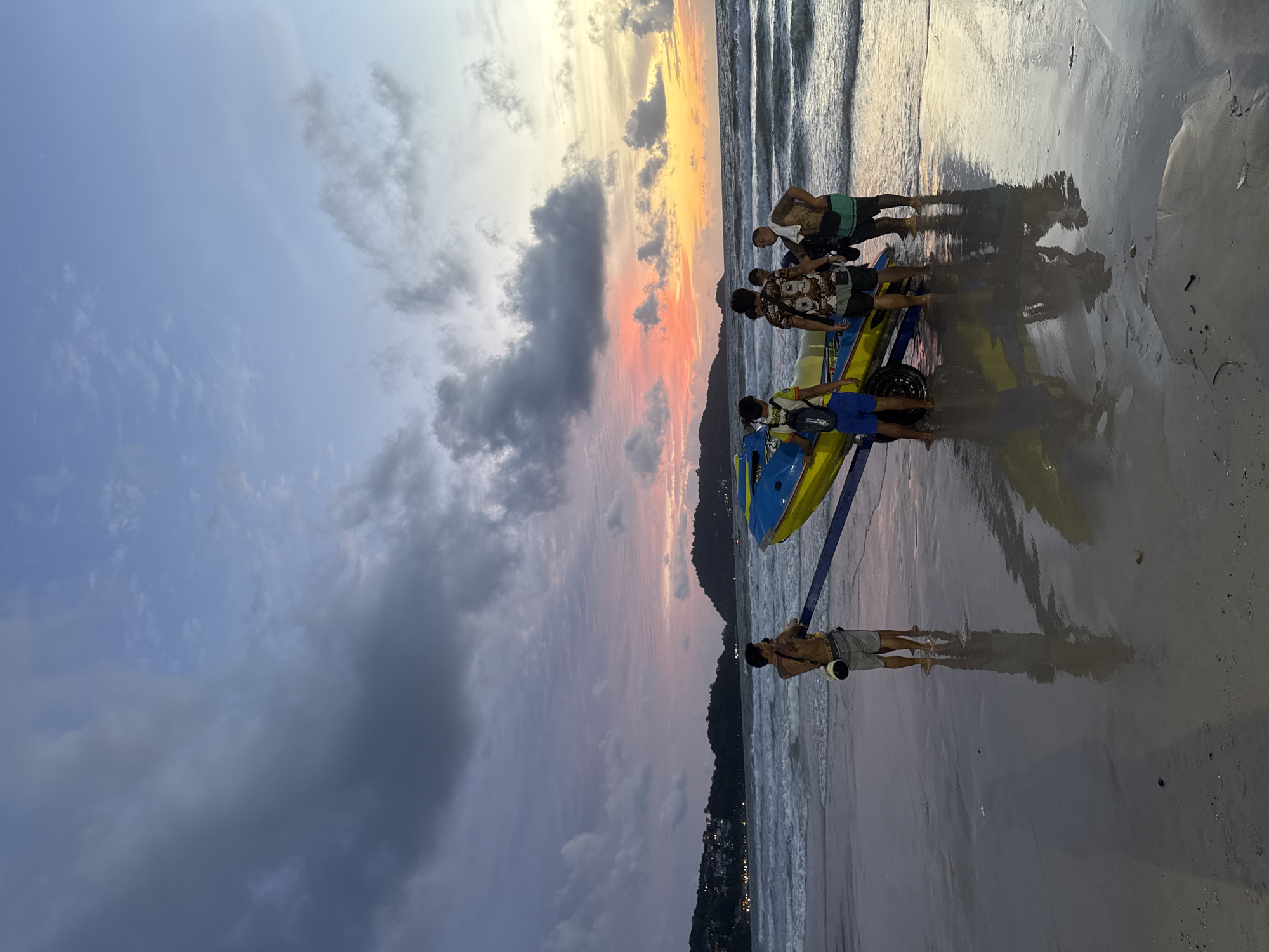 In Patong, Thailand, at dusk on a beach, a group of five individuals stands near a jet ski on a trailer. The sky displays a vibrant sunset with hues of orange, pink, and purple, juxtaposed with darker clouds. Waves gently wash ashore onto the wet sand. The group appears to be preparing to launch or retrieve the jet ski. The individual on the far left is wearing blue shorts and a backpack. The second person from the left has a patterned t-shirt and black shorts. The third person is wearing a t-shirt and dark shorts. The fourth person, with a towel draped over their shoulder, wears green shorts. The fifth person is mostly obscured by the group and the jet ski. In the background, a distant hillside is dotted with lights, suggesting the presence of buildings. The overall scene depicts a moment of leisure and activity on a popular Thai beach during sunset.