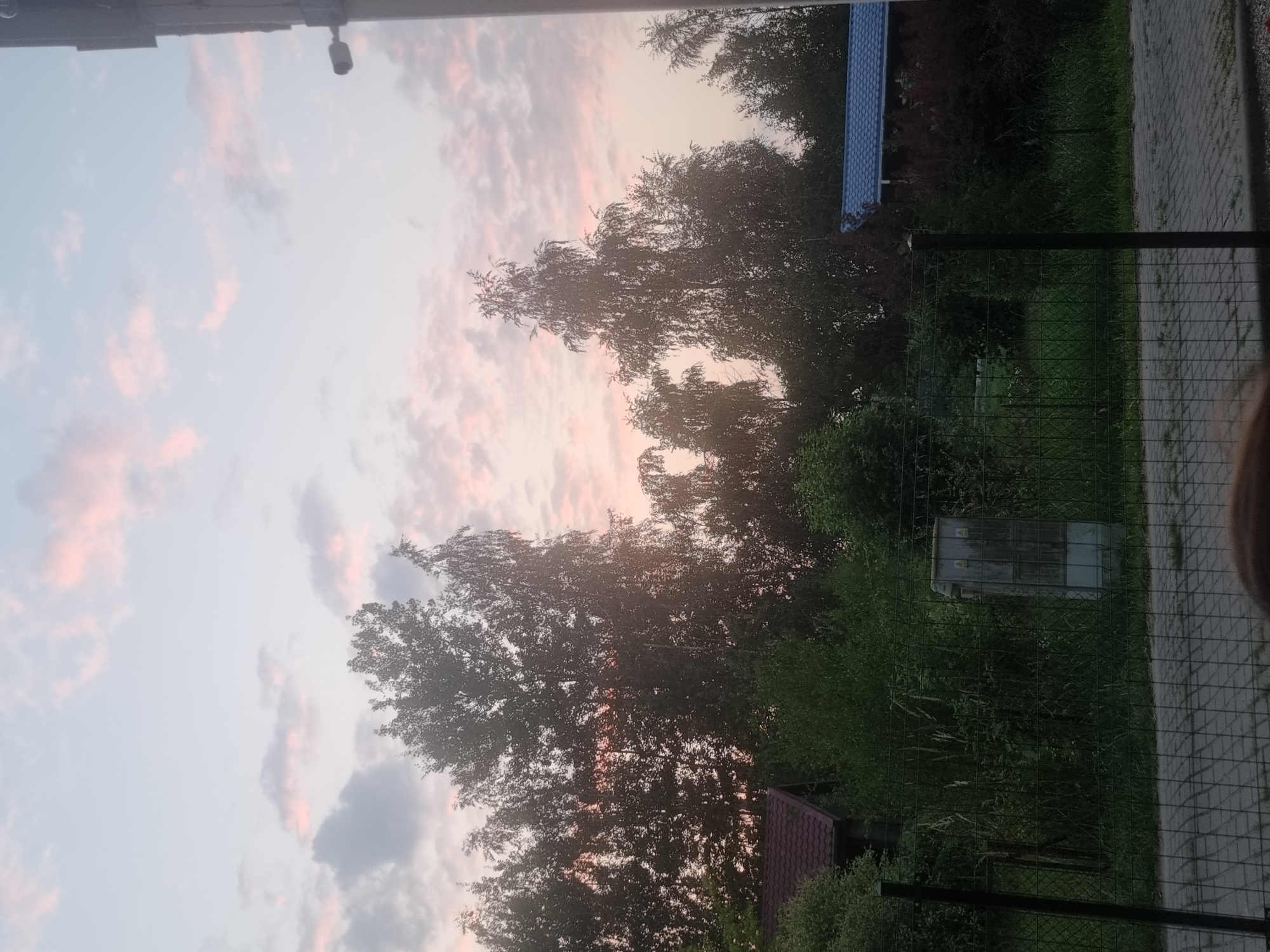 The image depicts an outdoor scene at dusk in the city of Szczaki, Poland, featuring a cloudy sky with pink hues. In the foreground, a wire fence runs horizontally across the frame, with a paved walkway visible beyond it. A utility box is situated behind the fence, amidst overgrown vegetation. Further back, a line of trees dominates the landscape, with a small building with a red roof on the left and another building with a blue roof on the right. A security camera is mounted on a vertical pole on the far right of the image. The overall impression is a serene, rural setting at the end of the day.