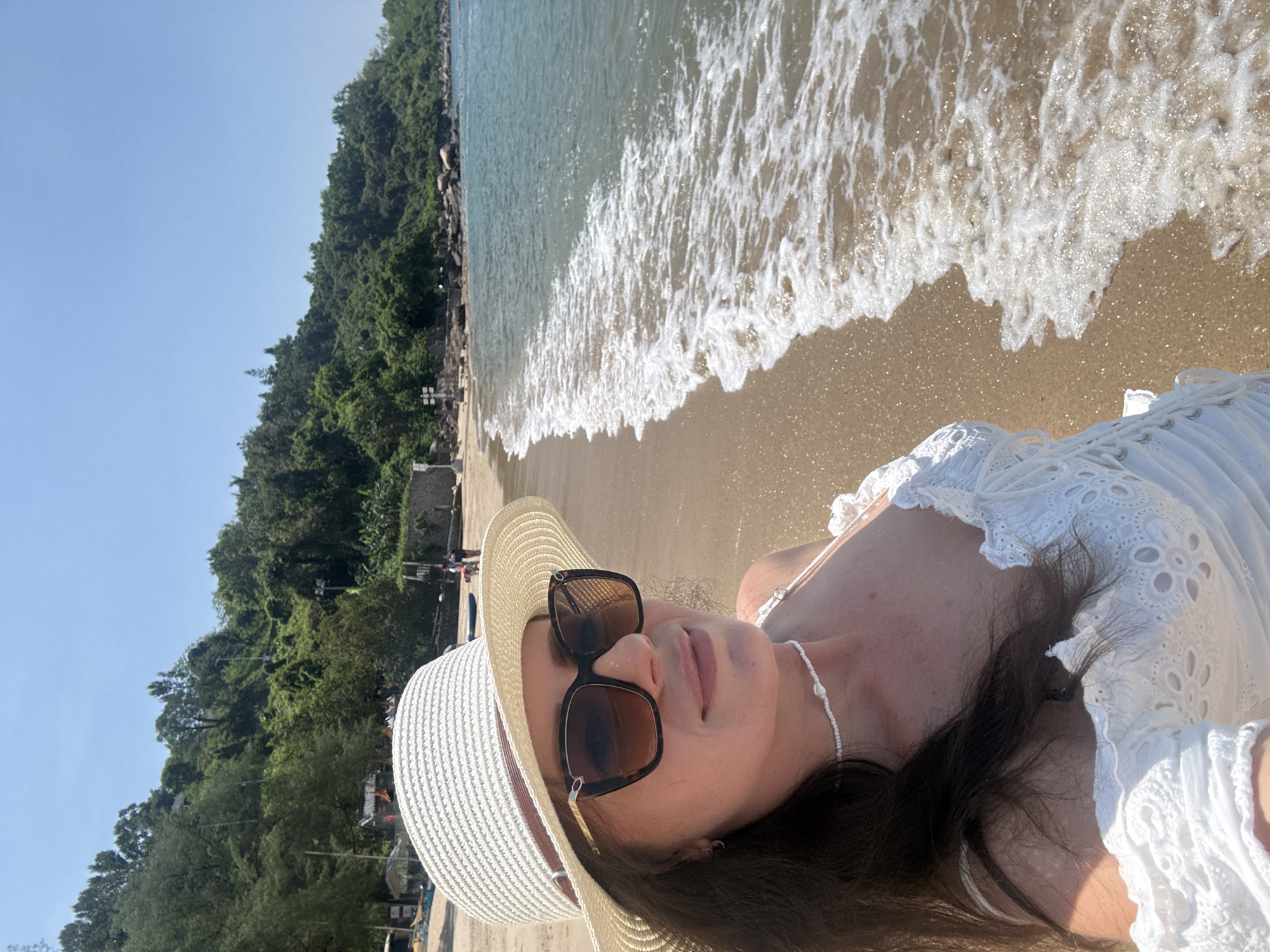 A woman is taking a selfie on a beach with lush green hills in the background. She is wearing a white off-the-shoulder dress, a straw hat, and sunglasses. The ocean waves are gently lapping at the shore. The scene appears to be in a tropical or subtropical coastal area, possibly in the city of Longgang District, China, given the presence of tropical vegetation and what appears to be a developed waterfront area with some structures.