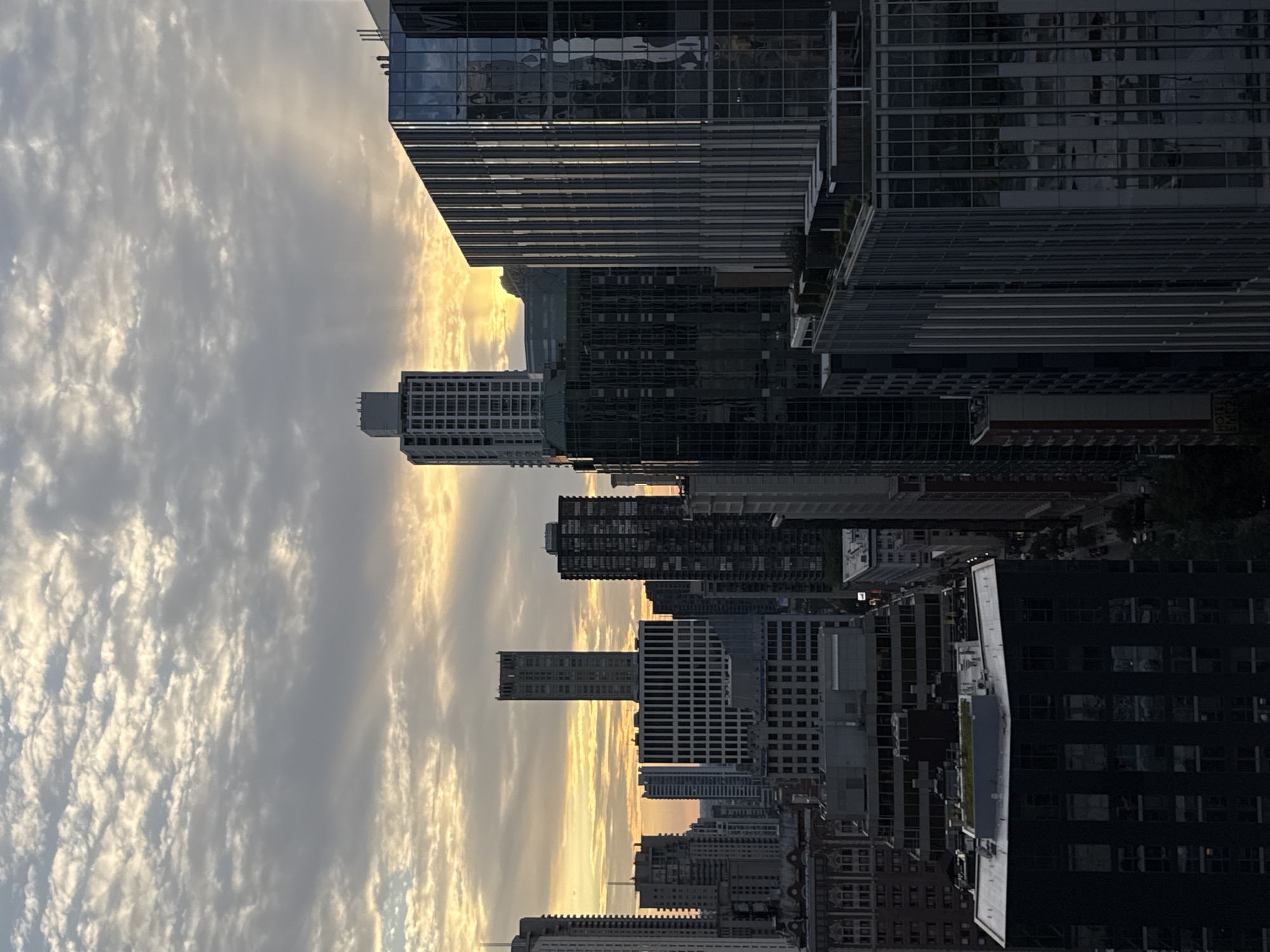 This is an aerial photograph of the Chicago skyline at sunset. The sun is low on the horizon, casting a warm glow over the city. The sky is filled with dramatic clouds, with rays of sunlight breaking through. In the foreground, several skyscrapers are visible, their glass facades reflecting the light of the setting sun. The buildings are of various heights and architectural styles, with some featuring sleek, modern designs and others displaying more traditional ornamentation. The scene is devoid of any people or vehicles, creating a sense of stillness and grandeur. The overall impression is one of urban beauty and the majestic power of nature.