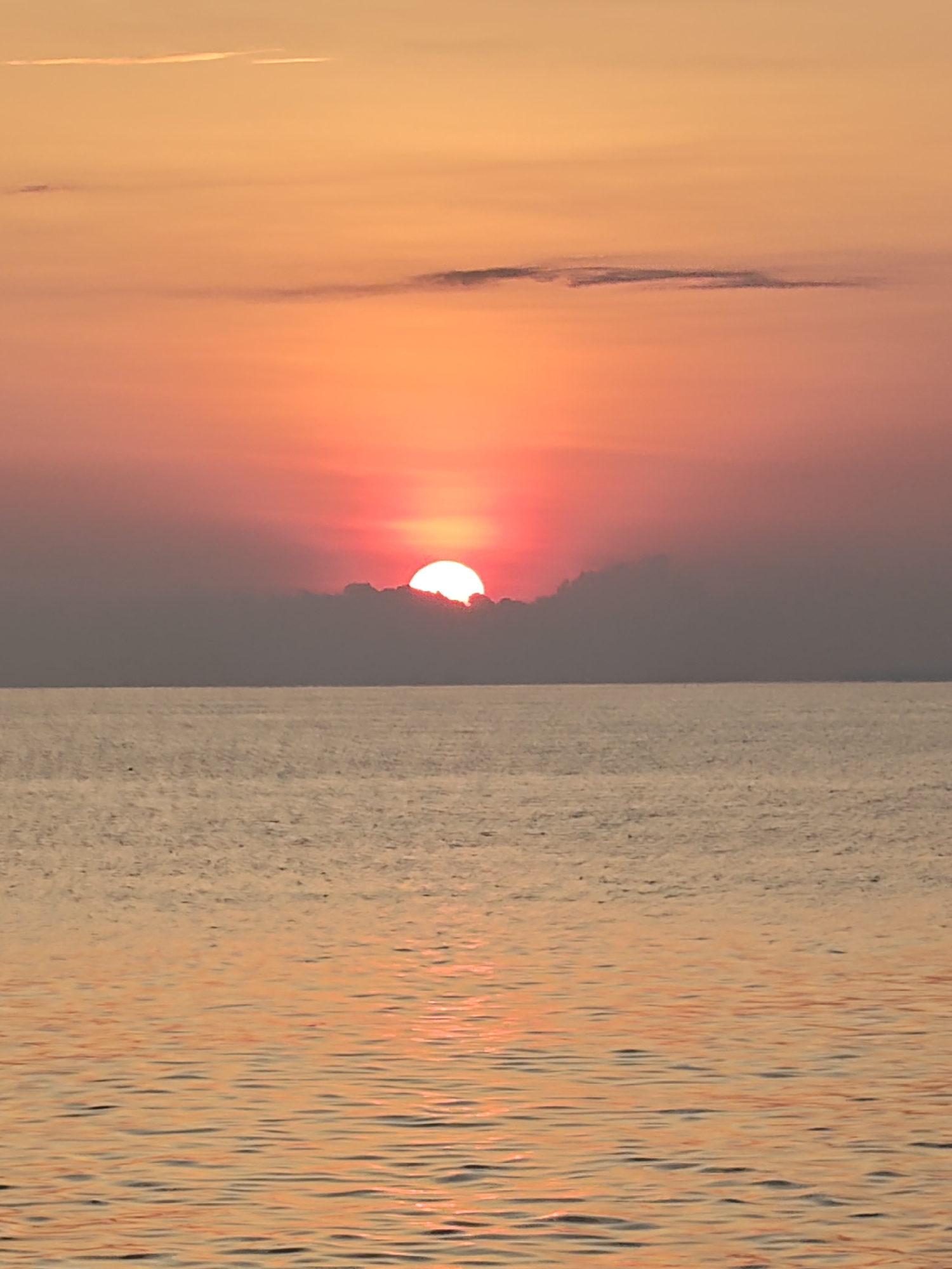 This image displays a sunrise or sunset over a large body of water, consistent with a coastal perspective from Lhokseumawe, Indonesia. The upper portion of the frame features a sky transitioning from a soft orange at the zenith to a vibrant red-orange nearer the horizon. A distinct, bright, red-orange sun is centrally positioned on the horizon line, with its lower half obscured by a dark, horizontal band of dense cloud cover. Several thin, elongated clouds are scattered across the upper orange sky. The lower half of the image depicts the water surface, characterized by subtle ripples that reflect the warm, orange-red hues of the sky and sun. No individuals, vessels, or specific terrestrial structures are present within the visible area.